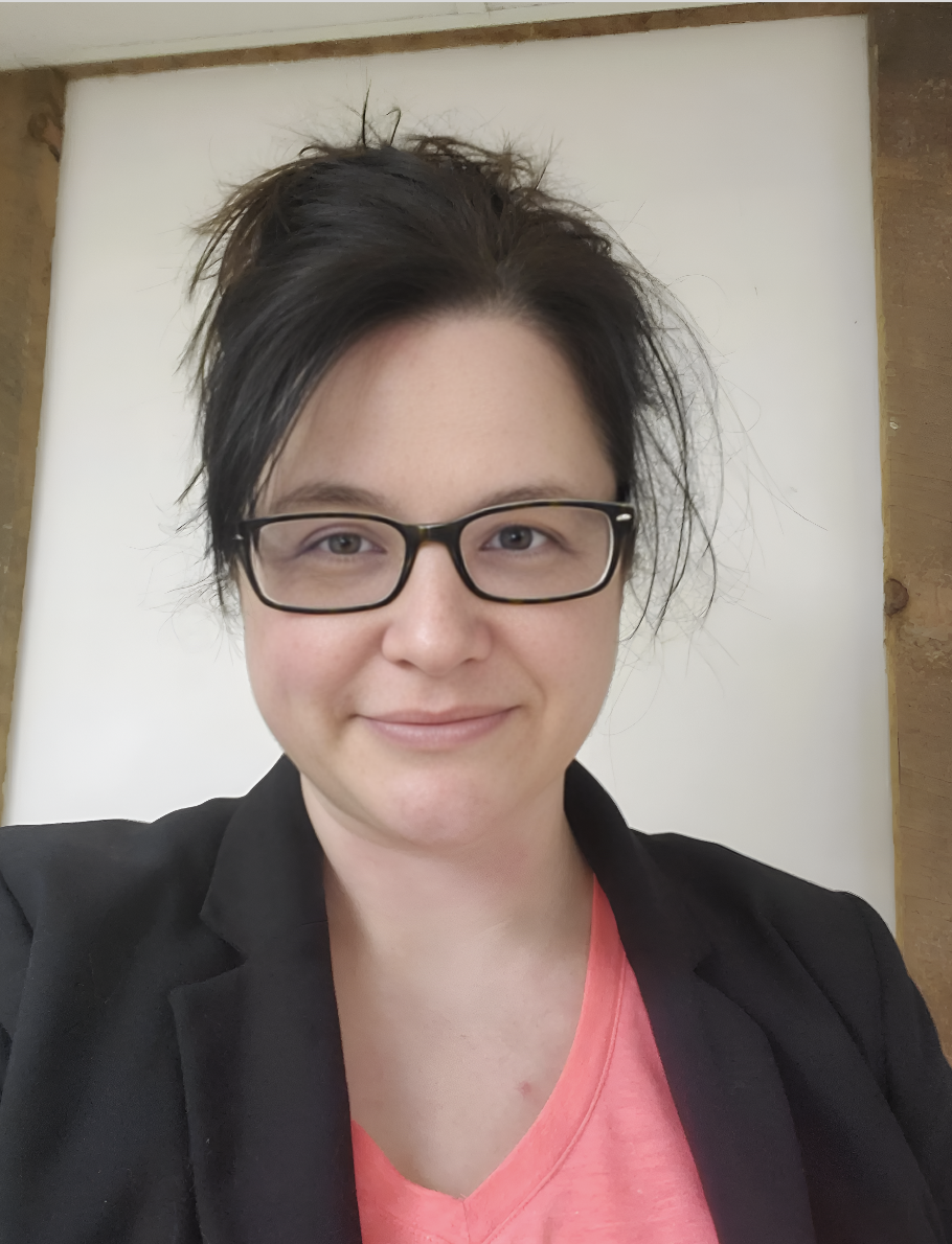 Smiling person with short, dark, slightly messy hair and glasses, wearing a black blazer and pink shirt, in front of a white background with wooden framing.