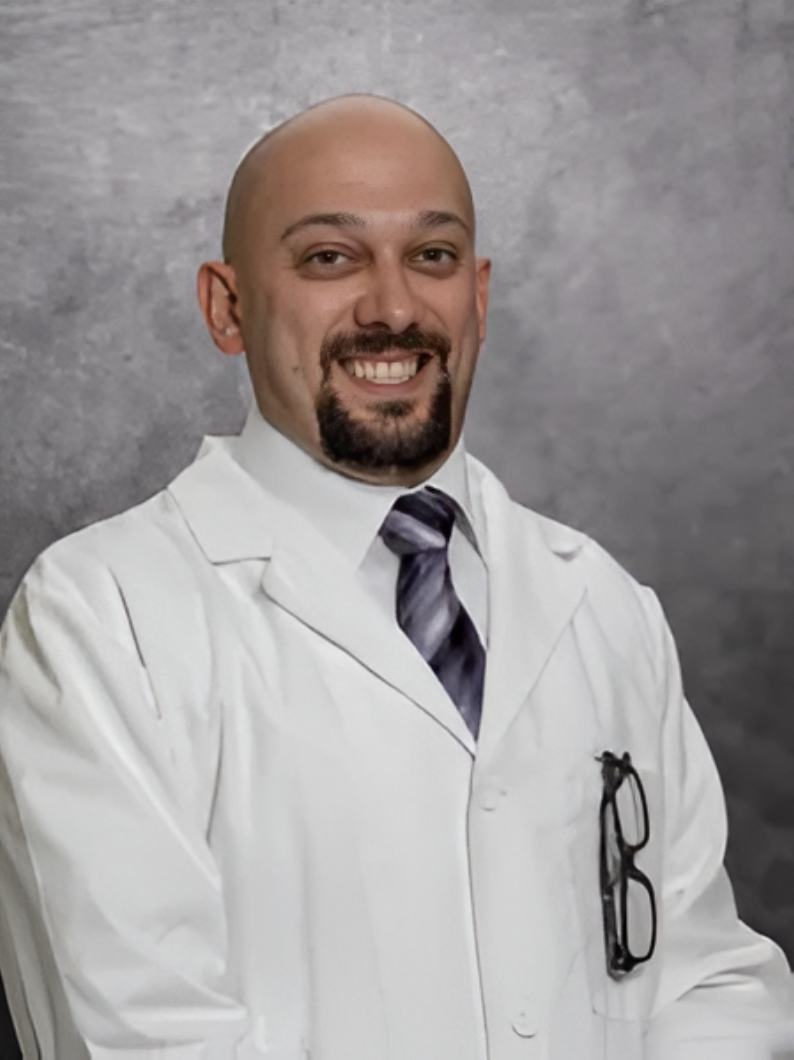 A smiling man in a white lab coat with a tie, standing against a gray background.