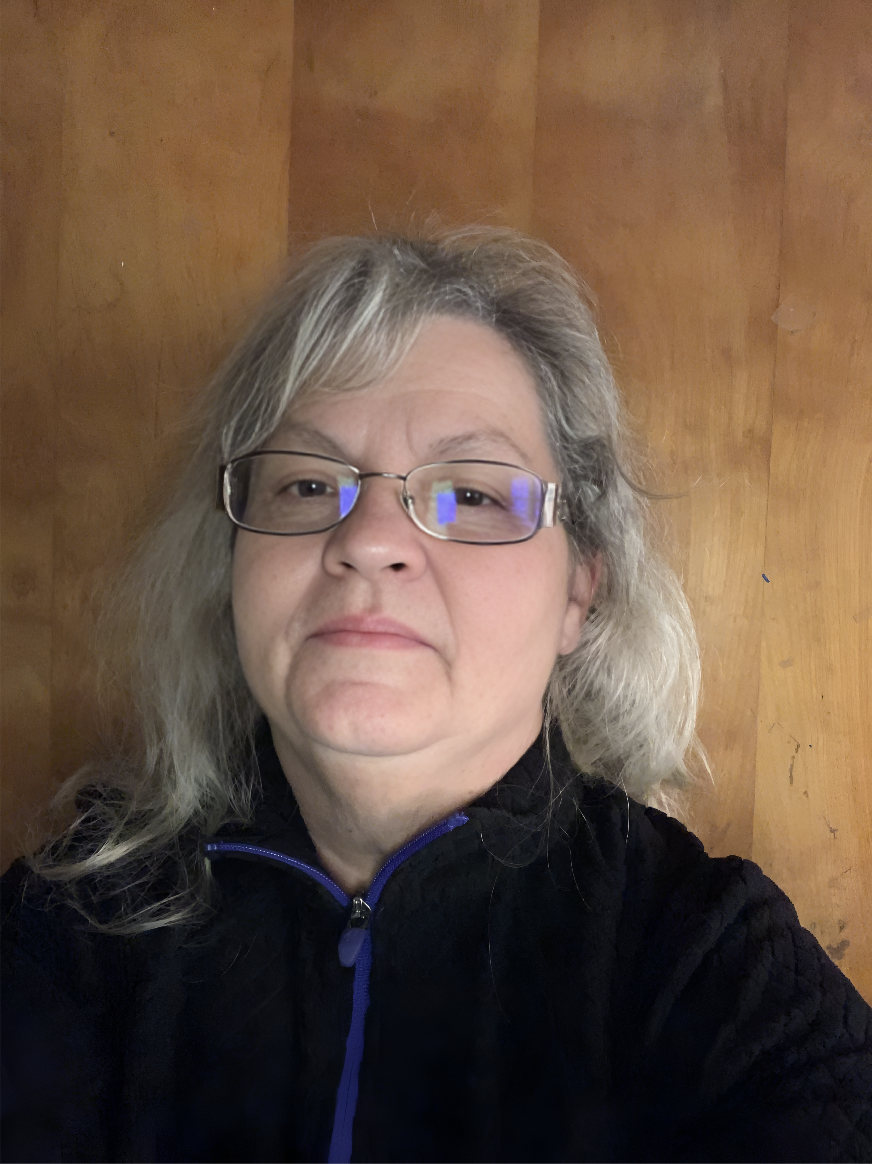 A woman with glasses and shoulder-length gray hair taking a selfie in front of a wooden wall, wearing a black jacket with a blue zipper.