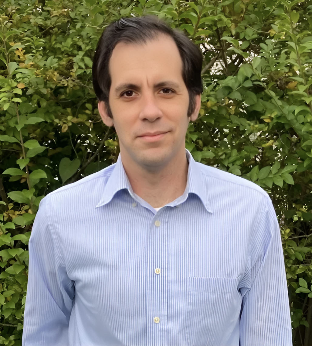 A man with dark hair and a slight smile, wearing a light blue, button-down shirt with thin white stripes, stands outdoors in front of green leafy bushes.
