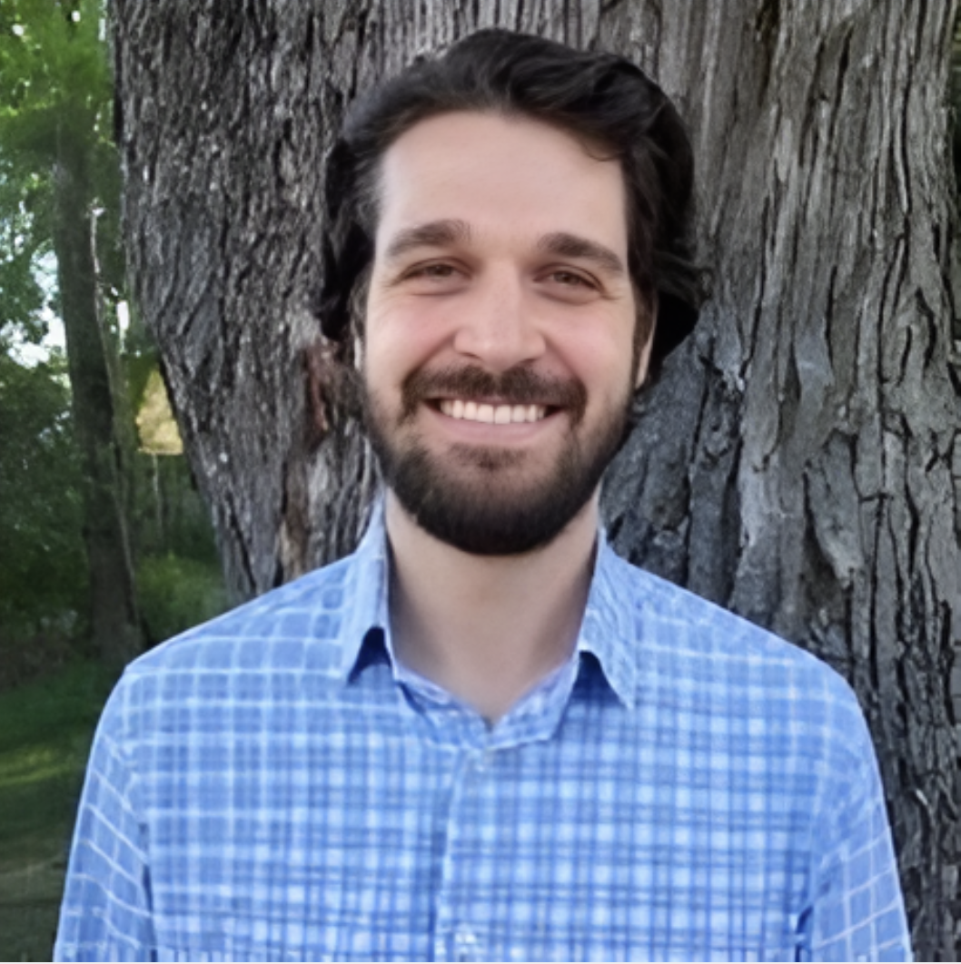 A man with dark hair and a beard, smiling, standing outdoors next to a large tree with green foliage in the background.