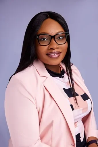 A woman with long black hair wearing glasses, a light pink blazer, and a patterned shirt, smiling at the camera against a purple background.