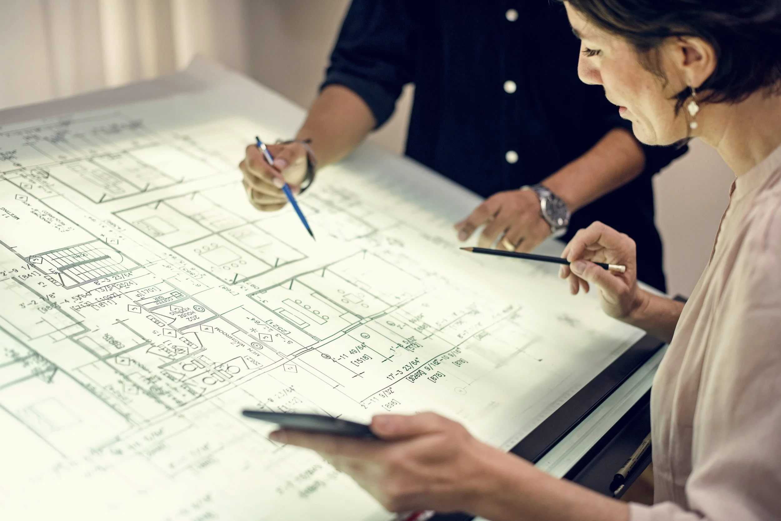 Two women looking at and discussing detailed architectural blueprints at a desk, one of them holding a smartphone.