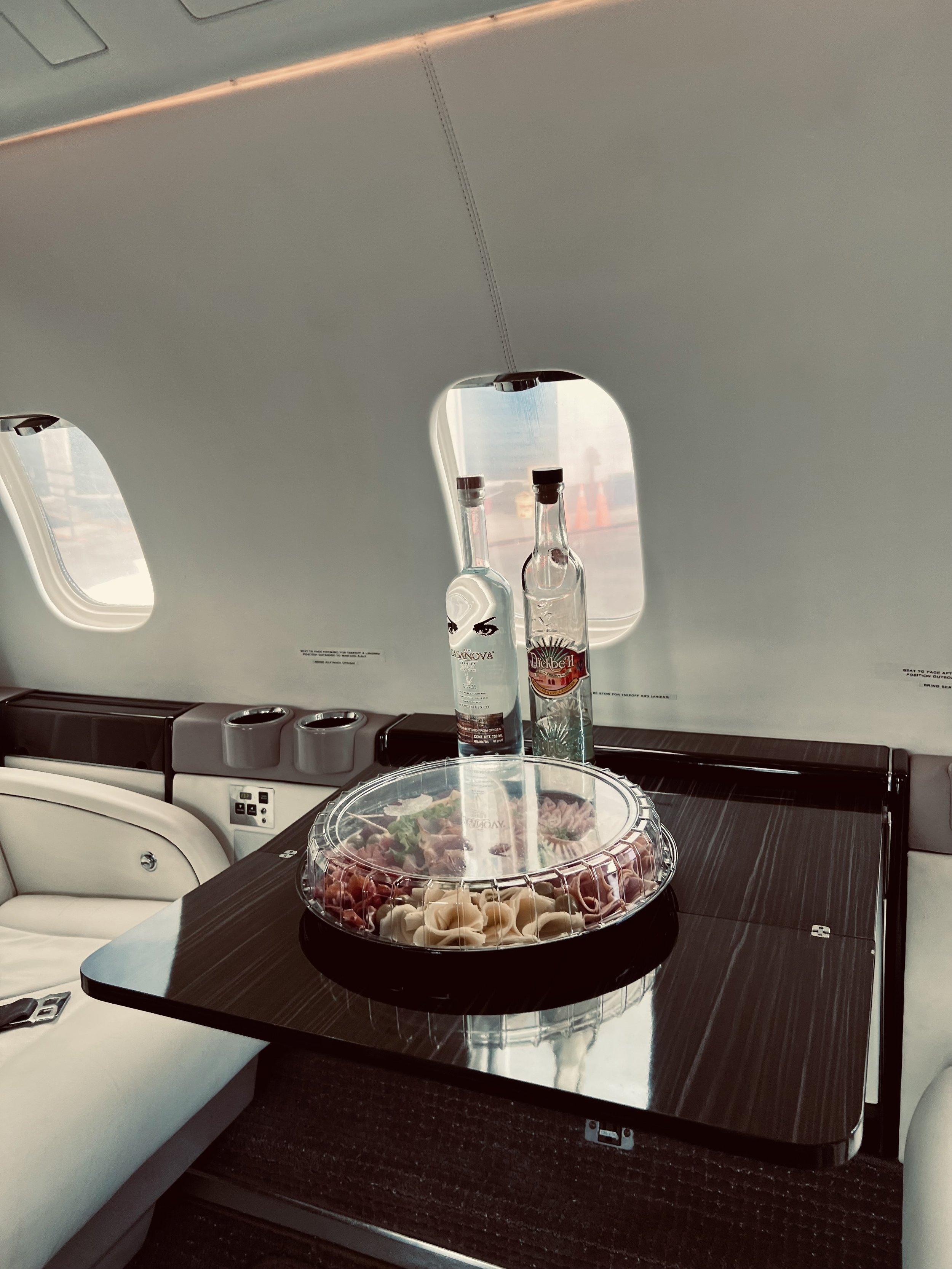 A tray of prosciutto and cheese on a table inside a private jet with two bottles of alcohol, one with a label, and the plane's windows in the background.