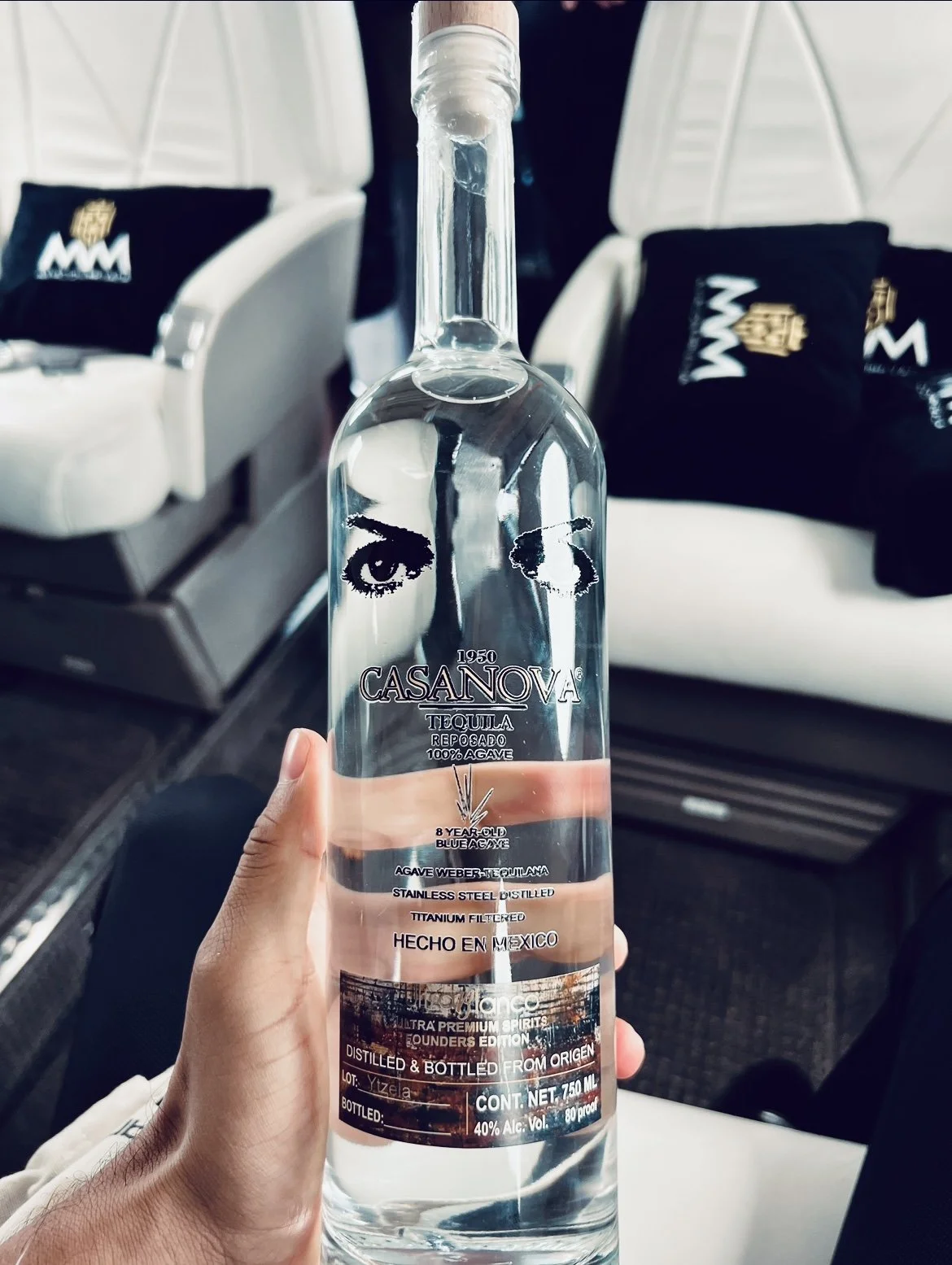 A hand holding a clear glass bottle of Casanova tequila with a design featuring eyes on the front. The bottle is inside an airplane with seats and black pillows in the background.