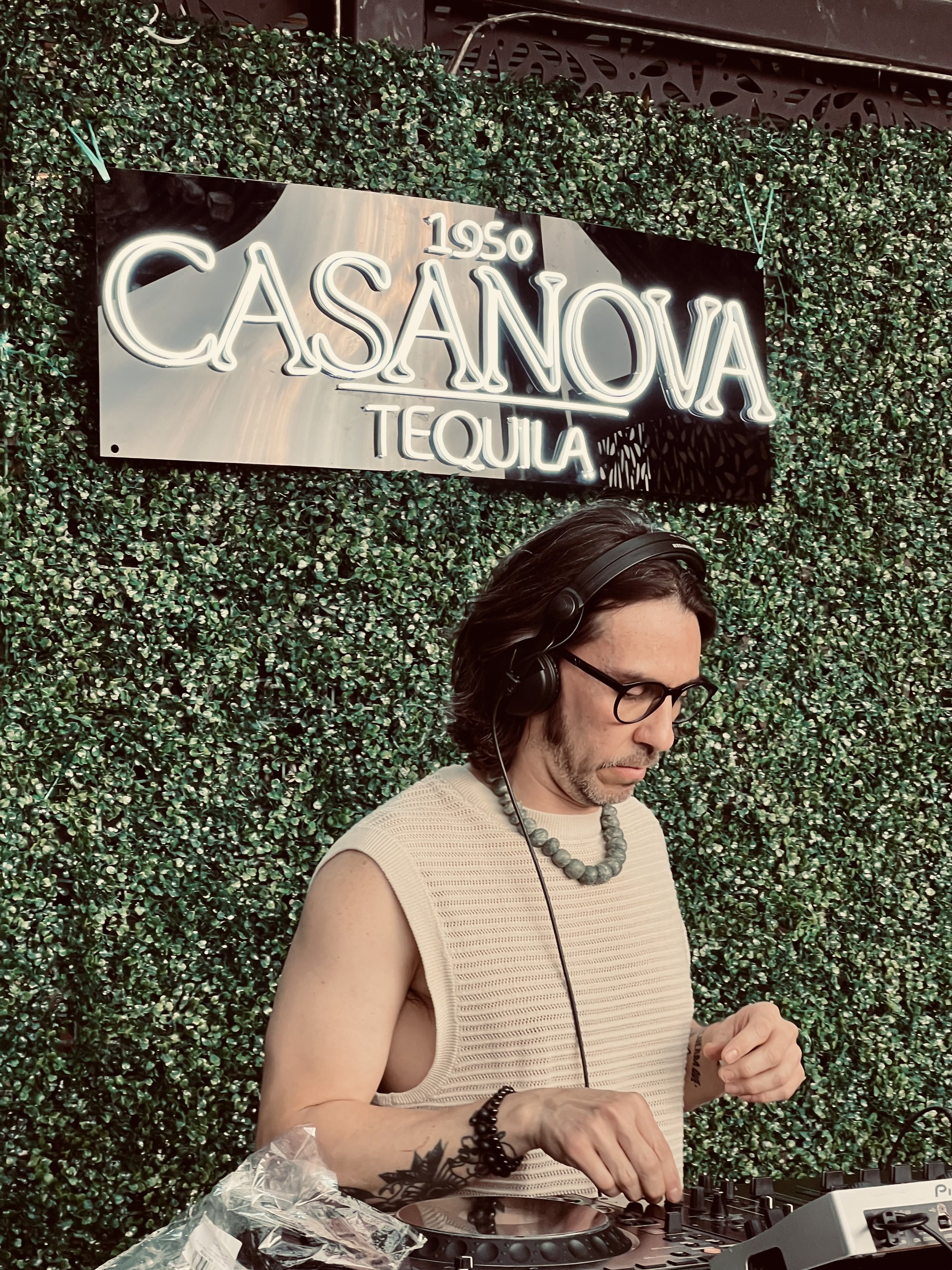 A man with long dark hair, glasses, and a beard, wearing a sleeveless beige top, is DJing with headphones on, against a green hedge wall with a neon sign that reads '1950 Casanova Tequila.'