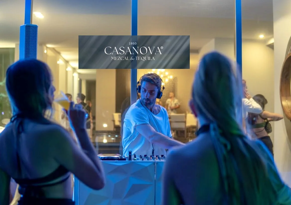 DJ playing music at a party with two women drinking cocktails in the foreground, inside a modern venue with a sign that reads 'Casanova Mezcal & Tequila'.