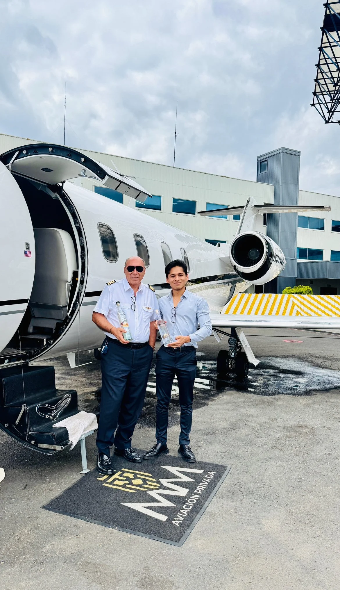 Two men standing in front of a private jet, holding bottles of alcohol, on the tarmac. One man is in a pilot uniform and the other in business attire. The jet has open doors and is parked near a building with the logo 'AVIANCA.'