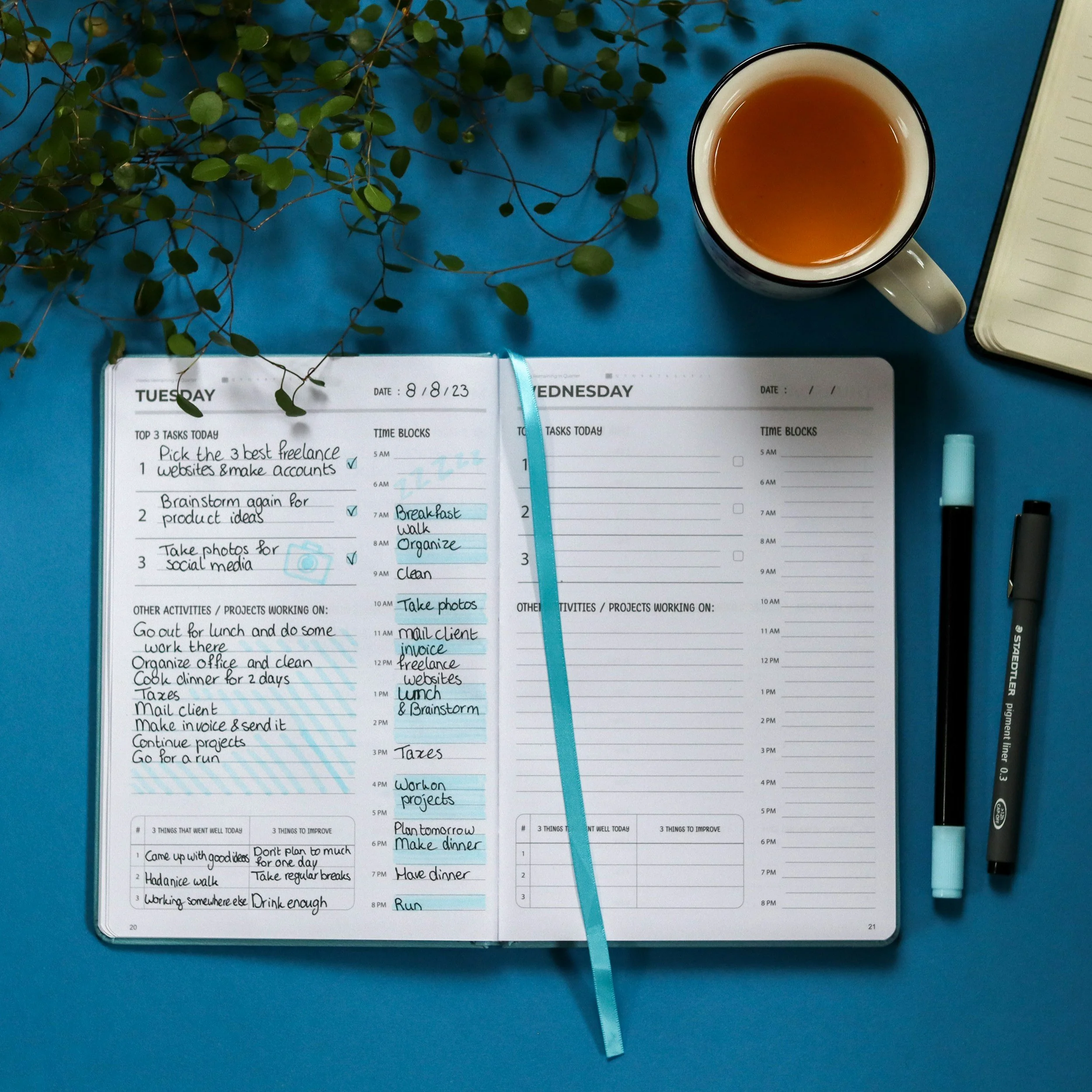 Open planner for Tuesday, August 18, 2023, with tasks and activities listed, a cup of tea, a pen, and a notebook on a blue desk, with green plant leaves in the top left corner.