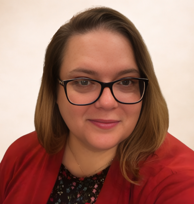 A woman with shoulder-length brown hair, glasses, and a slight smile, wearing a red jacket over a floral top, standing against a plain light-colored background.