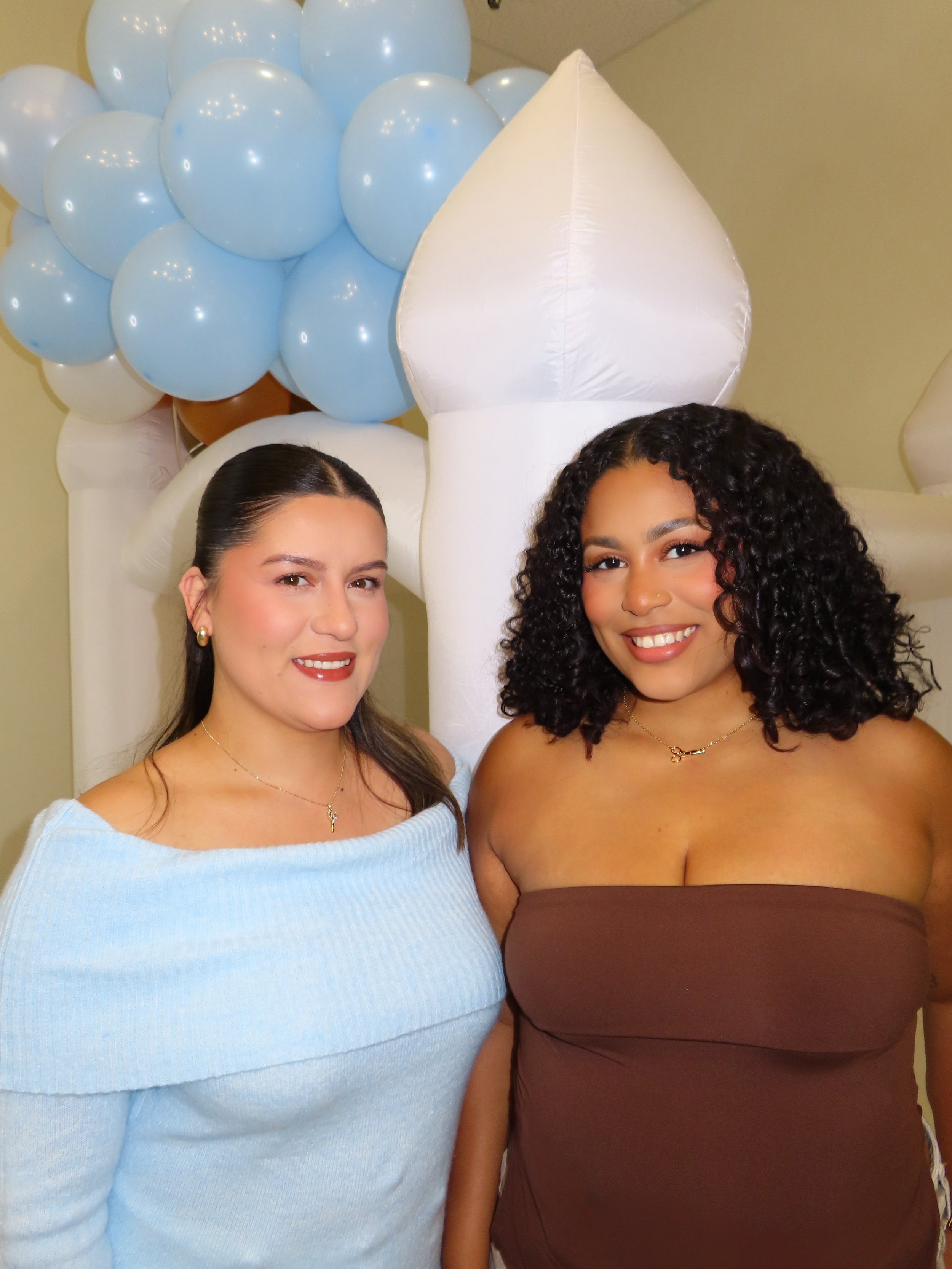 Two women smiling at a party, with blue and white balloons and large white balloon figures in the background.