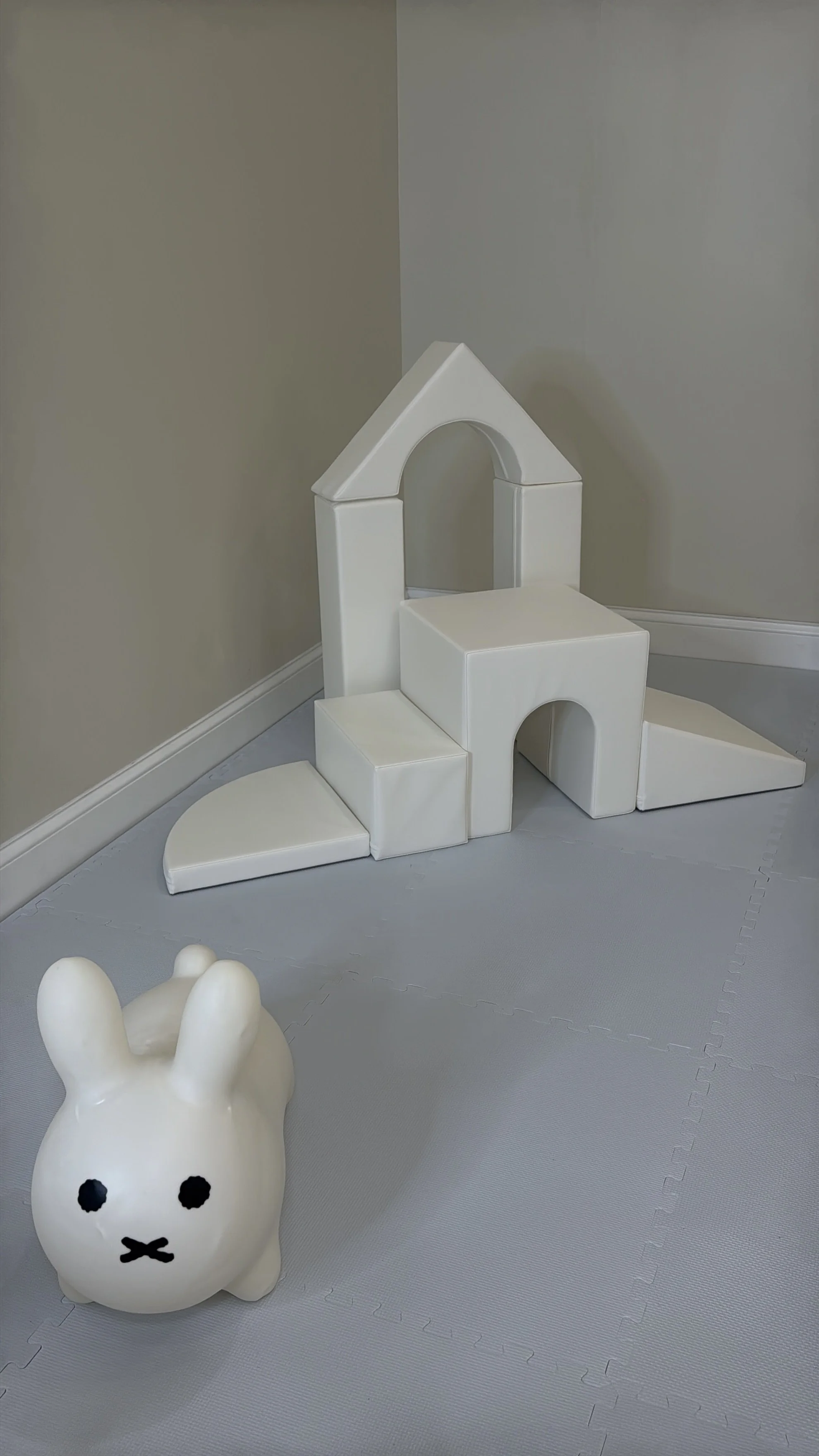 A white bunny-shaped stool and soft play equipment in the shape of a house with steps and arches, set against a plain wall and floor.