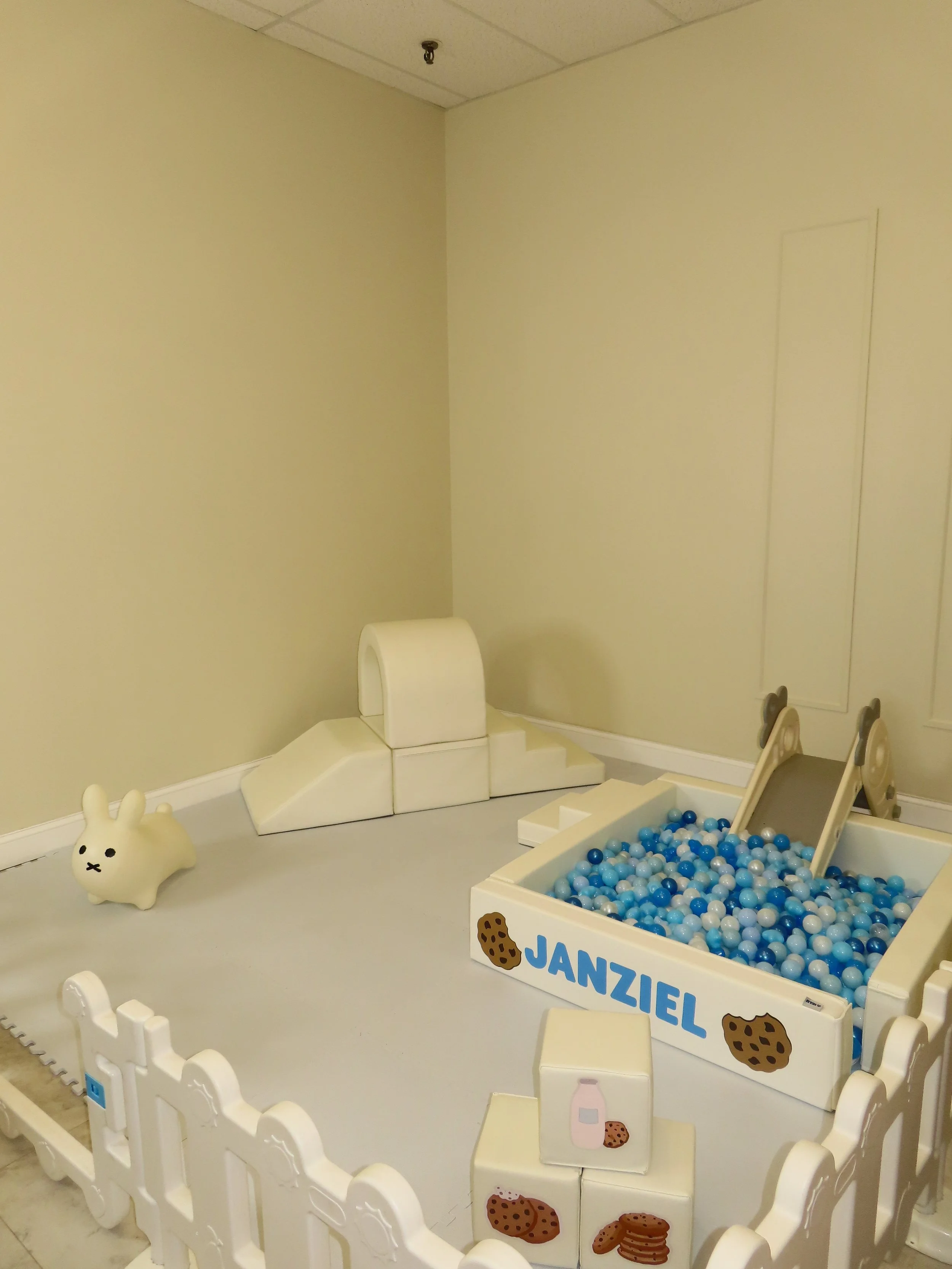 Children's playroom with pastel-colored toys, a ball pit labeled 'Janziel' with blue, white, and gray balls, a small slide, a bunny-shaped toy, and soft foam blocks with cookie and milk prints.