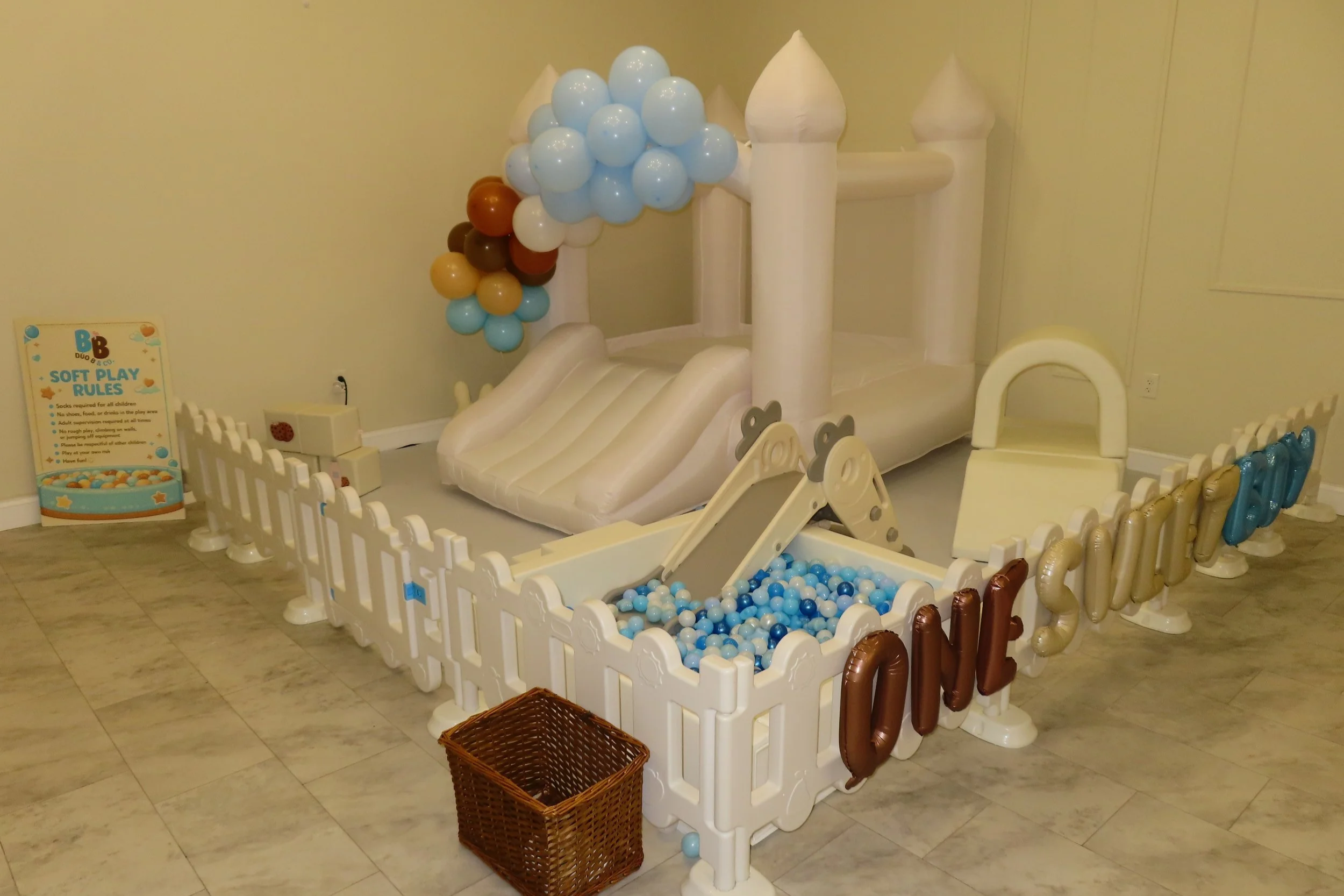 Inflatable indoor playground designed to resemble a castle with a slide, ball pit, and fencing, decorated with balloons, and a small sign with rules.