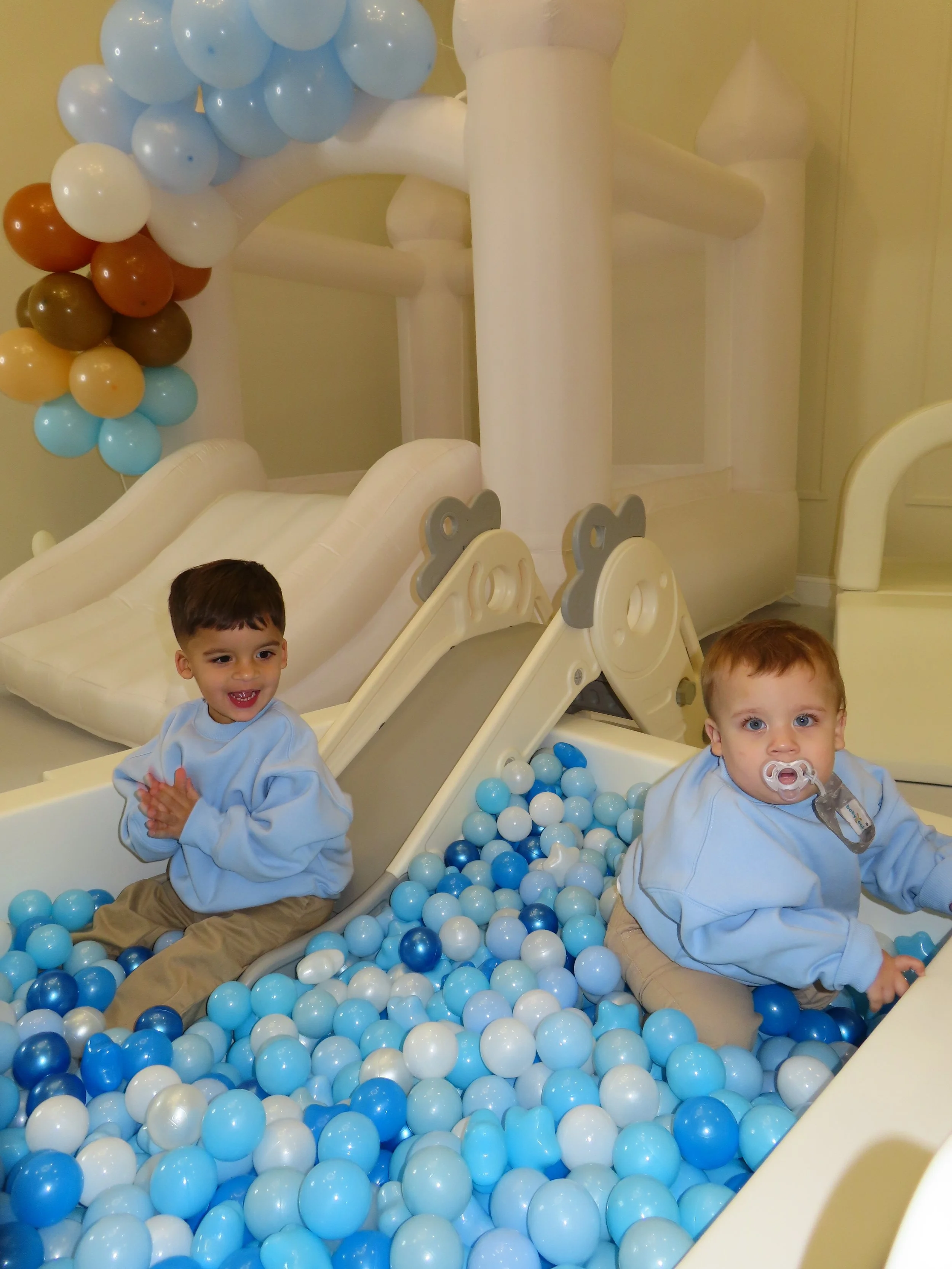 Two young children playing in a ball pit with a slide, surrounded by pastel blue, white, and brown balloons.