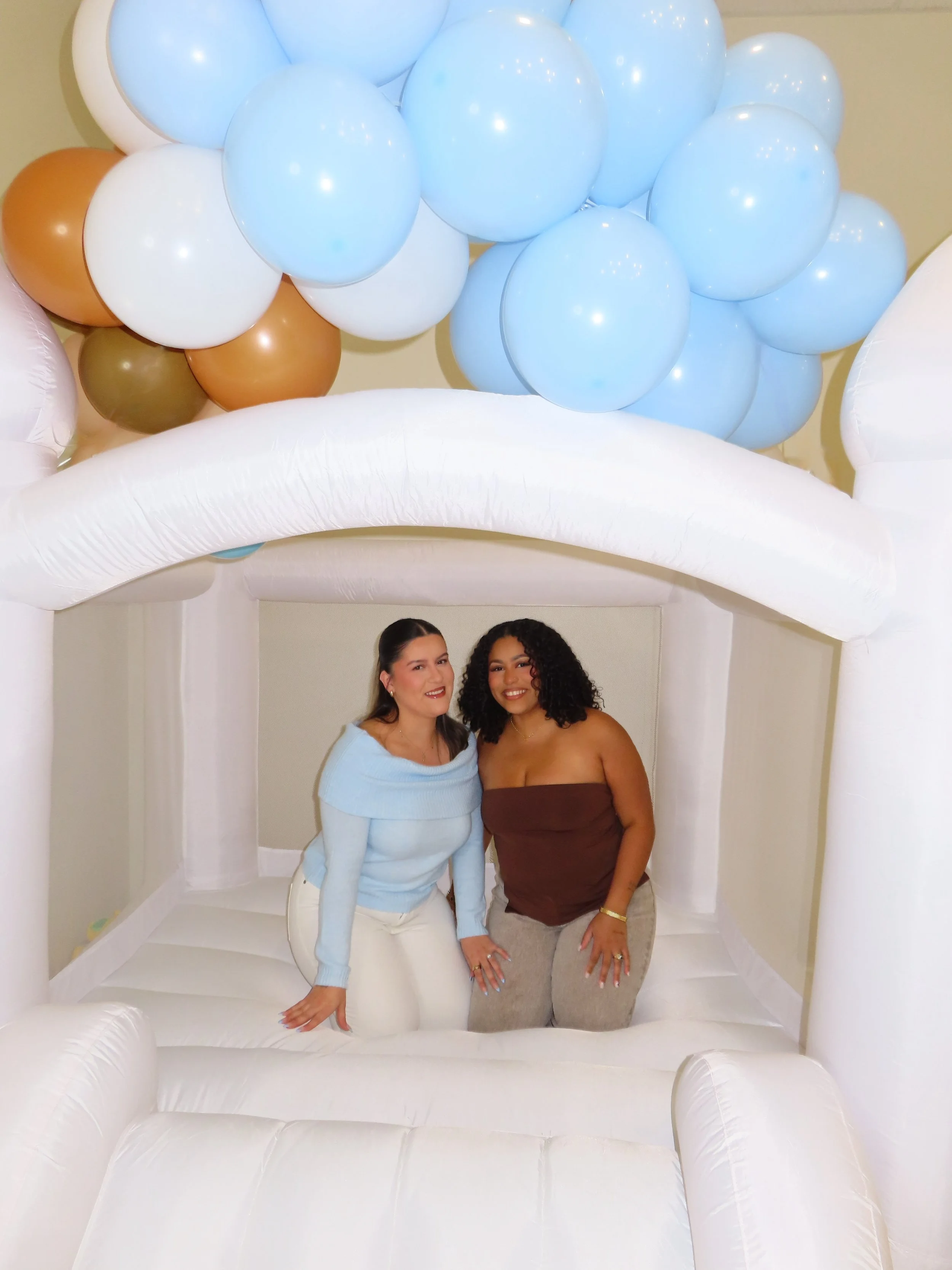 Two women kneeling inside an inflatable cloud-shaped structure with blue, white, brown, and olive green balloons floating above.