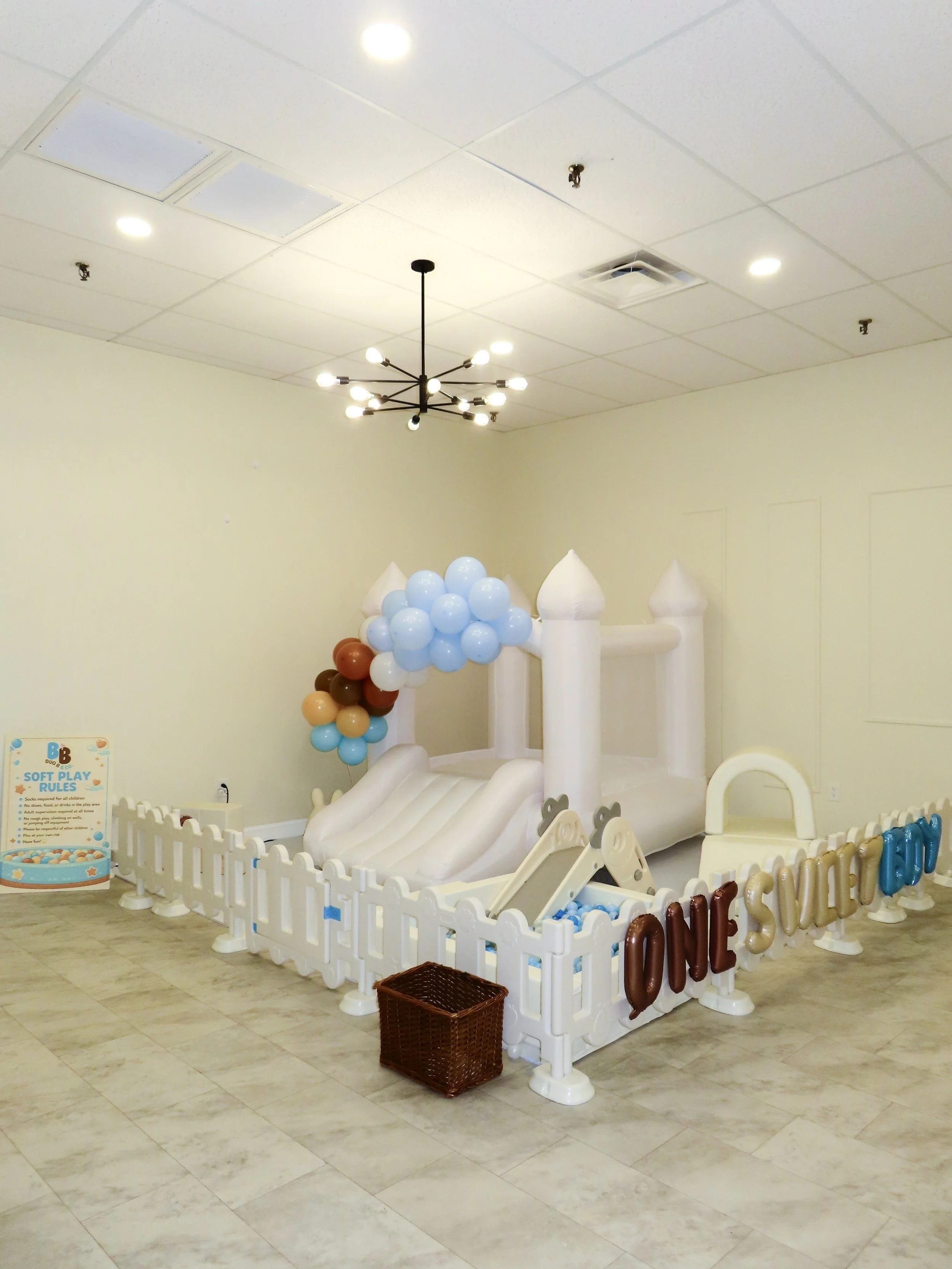 Inflatable castle with slide, decorated with balloons, enclosed by a white fence, and a sign celebrating a child's first birthday.