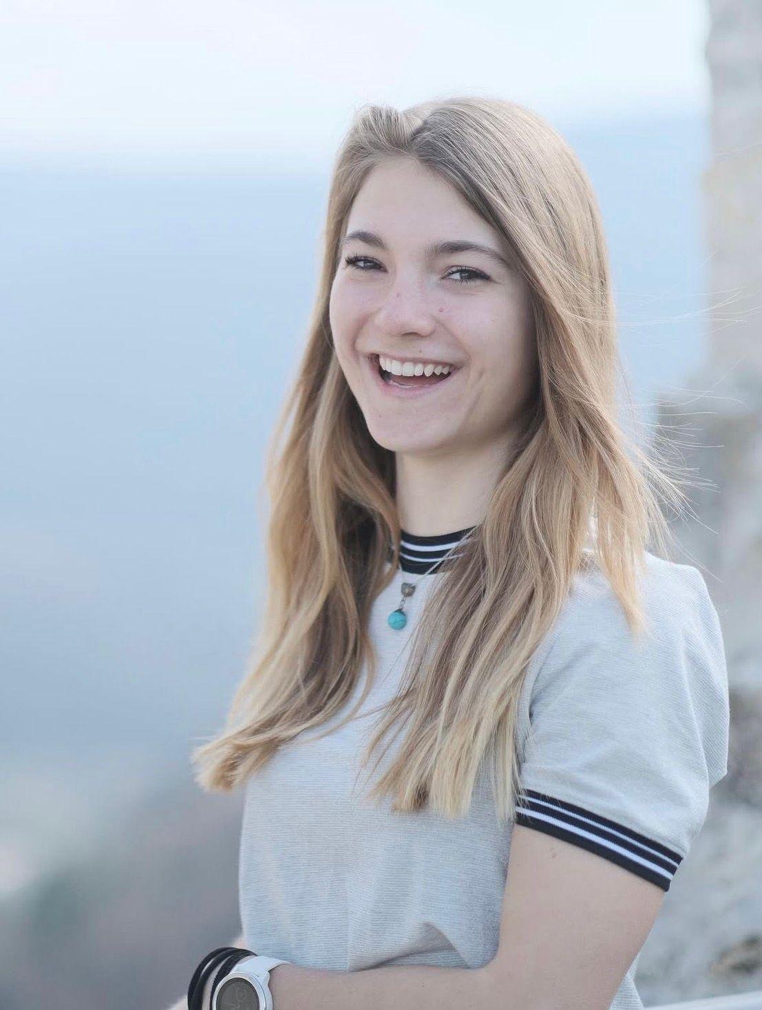 Lächelnde junge Frau mit langem, blondem Haar, trägt ein weißes T-Shirt mit schwarzen, weißen Streifen an den Ärmeln und Hals, und einen schwarzen Armband samt Smartwatch, vermutlich am Strand oder in der Nähe des Wassers.