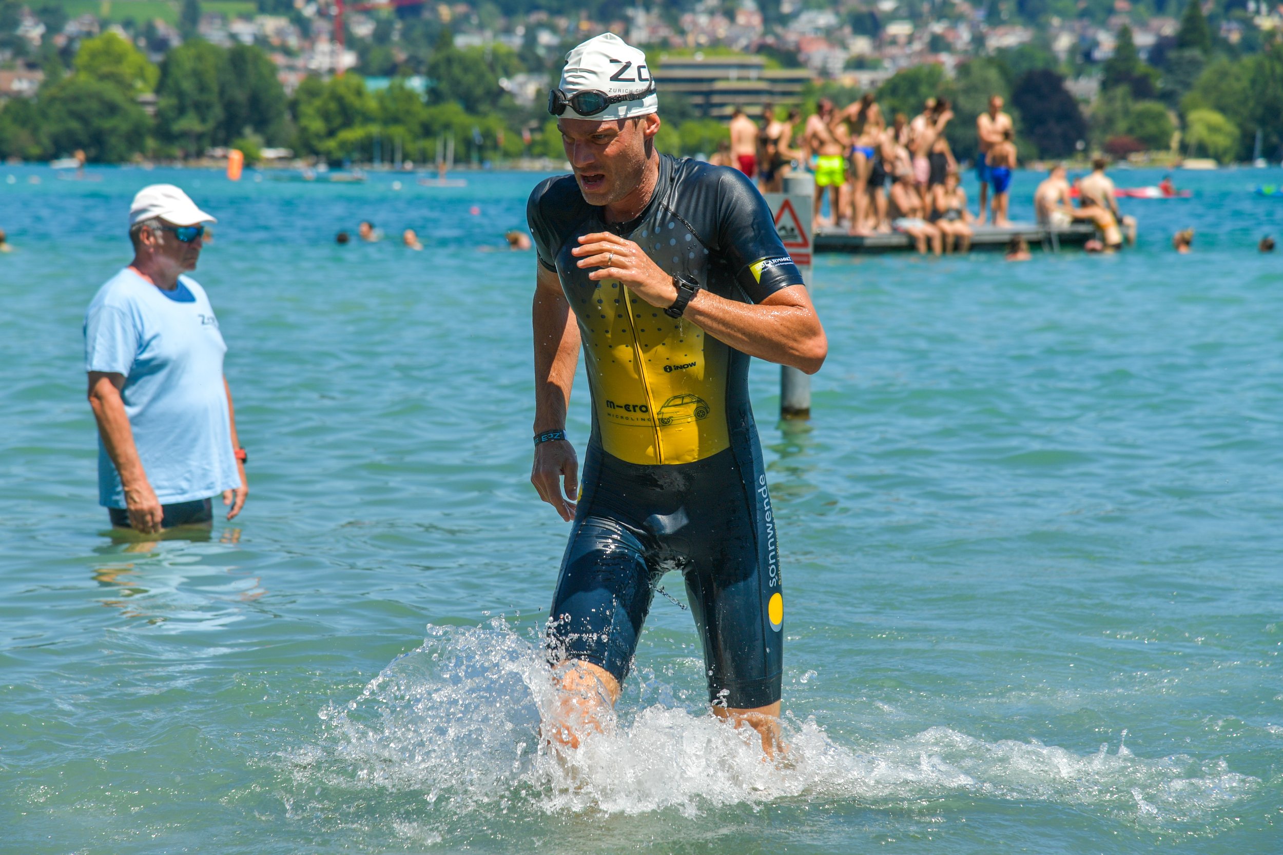 Ein männlicher Athlet beim Triathlon, schwimmend im See, trägt eine Neoprenanzug, Schwimmbrille und eine Badekappe, während im Hintergrund Zuschauer auf einem Boot sitzen.