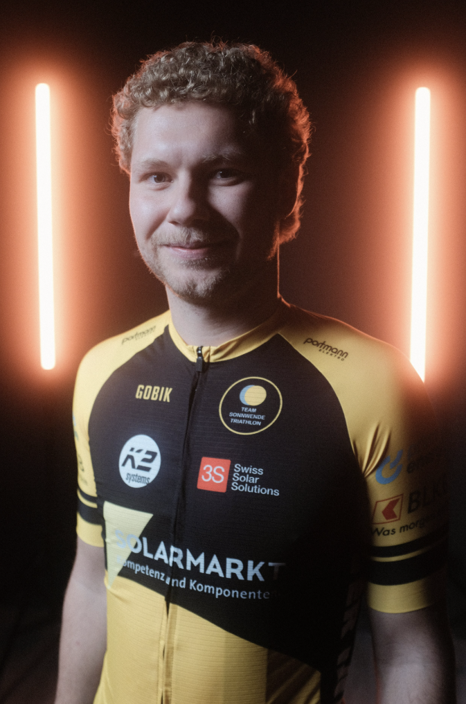 Porträt eines männlichen Radfahrers in einem schwarzen und gelben Radtrikot vor einem dunklen Hintergrund mit orangefarbenen Lichtern.