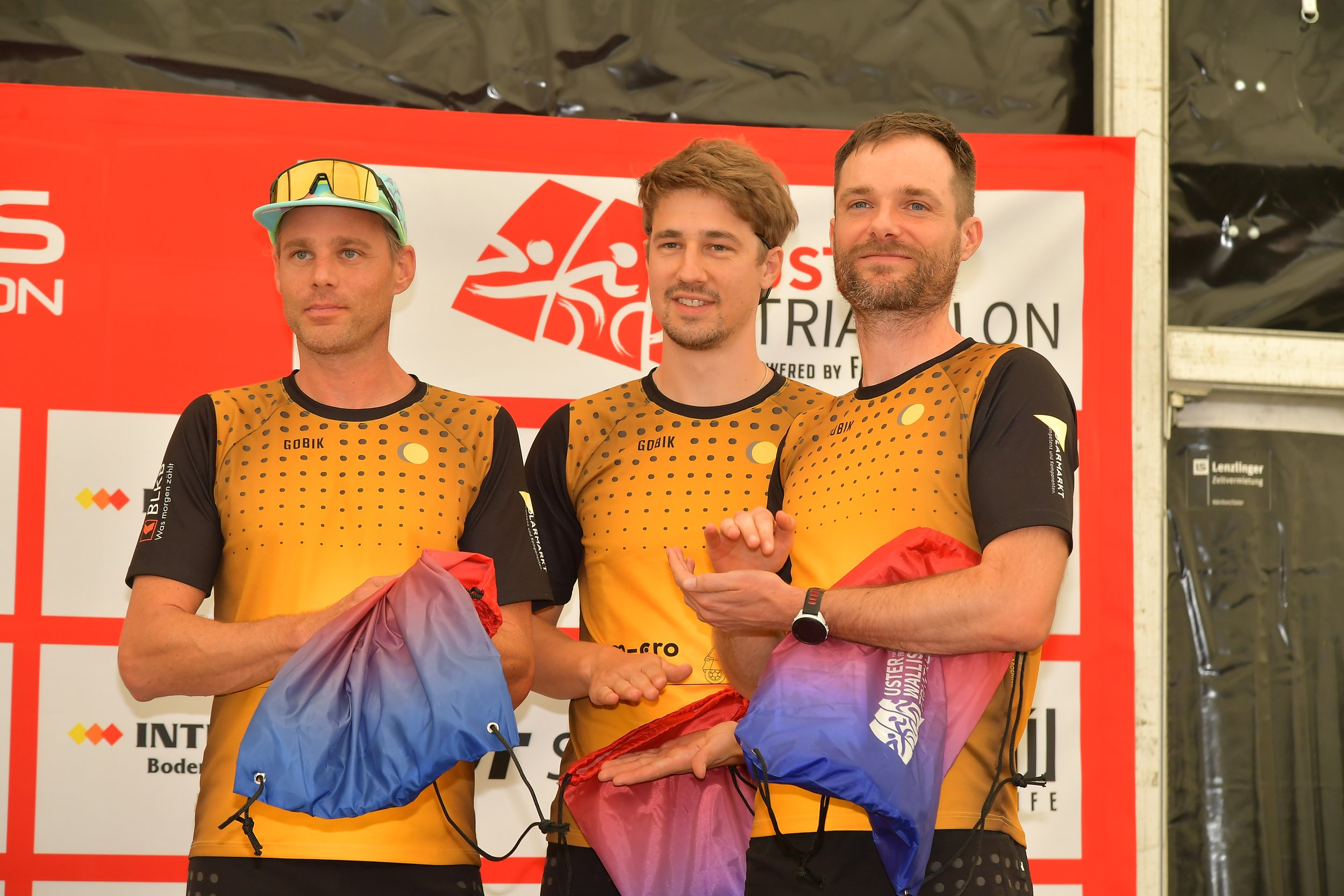 Drei Männer in gelben und schwarzen Triathlon-Trikots stehen auf einem Podium nach einem Wettkampf. Sie halten Auszeichnungstaschen in den Händen, während im Hintergrund ein rotes Banner mit Logos und Text hängt.