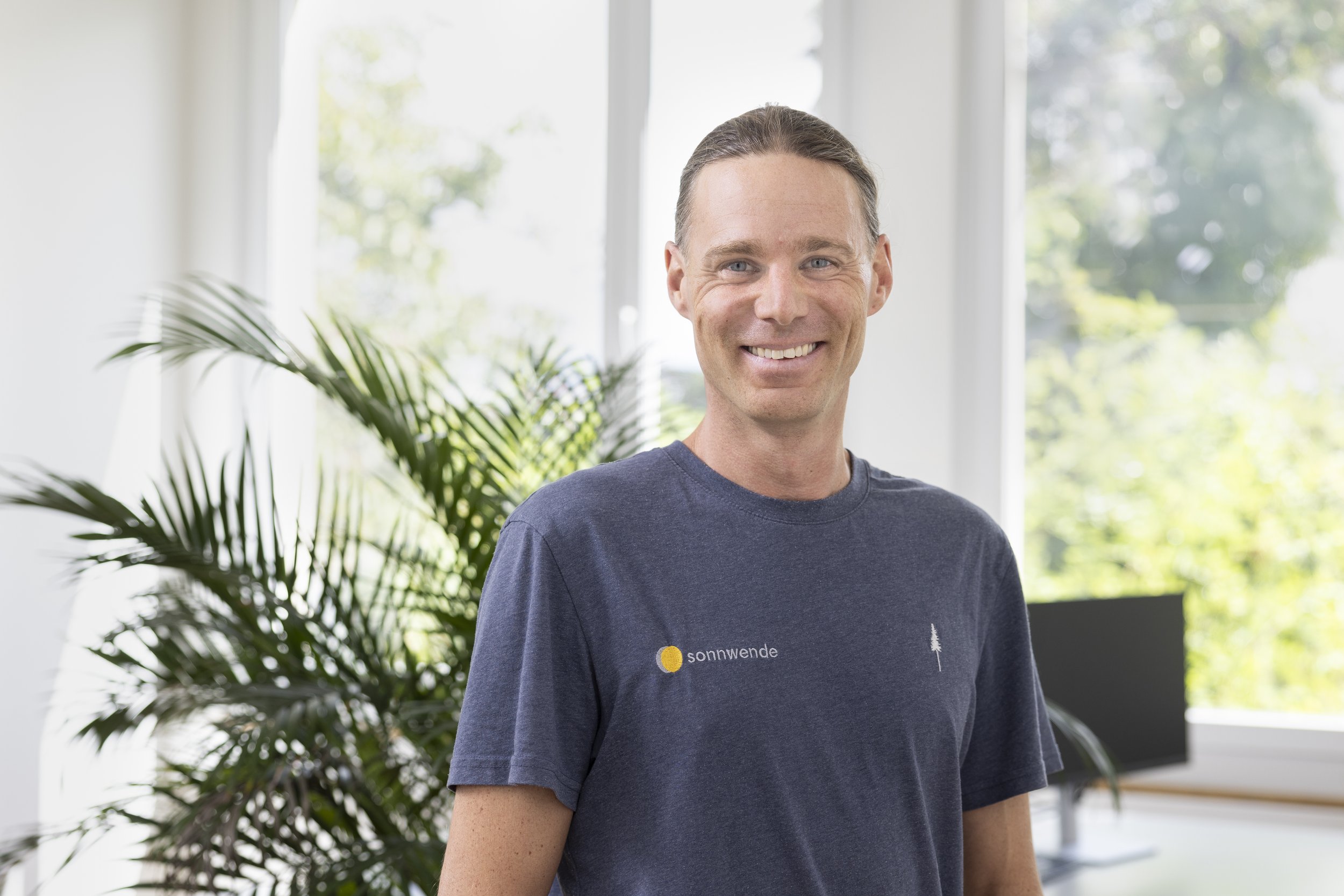 Lächelnder Mann in einem blauen T-Shirt mit Aufdruck 'sonnwende' steht in einem hellen Raum mit Pflanzen und einem Computerbildschirm im Hintergrund.