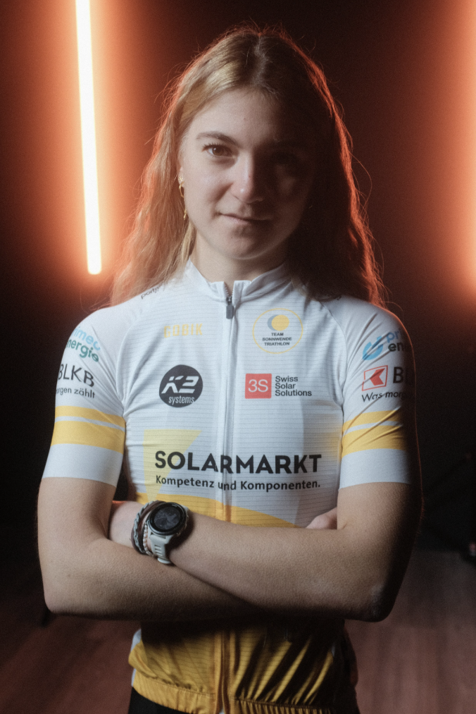 Junge Outdoor-Sportlerin in weißer Sonnenenergie-Lizenz-Jersey mit Sponsorenlogos, braune Haare, dunkle Hintergrundbeleuchtung mit warmem Licht, trägt eine Sportuhr am Handgelenk.