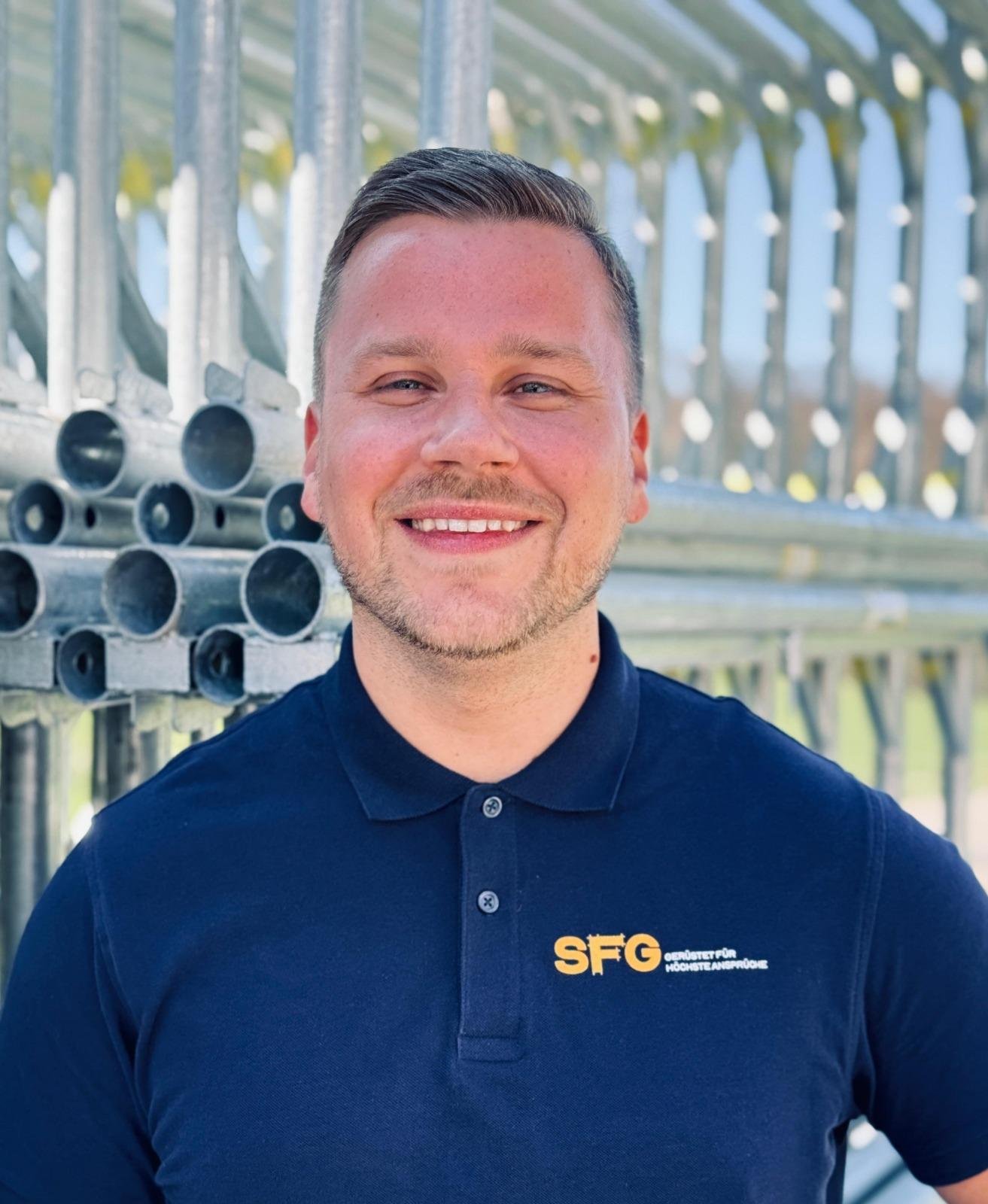 Mann trägt schwarze Poloshose mit SFG Logo, steht vor einer Metallkonstruktion