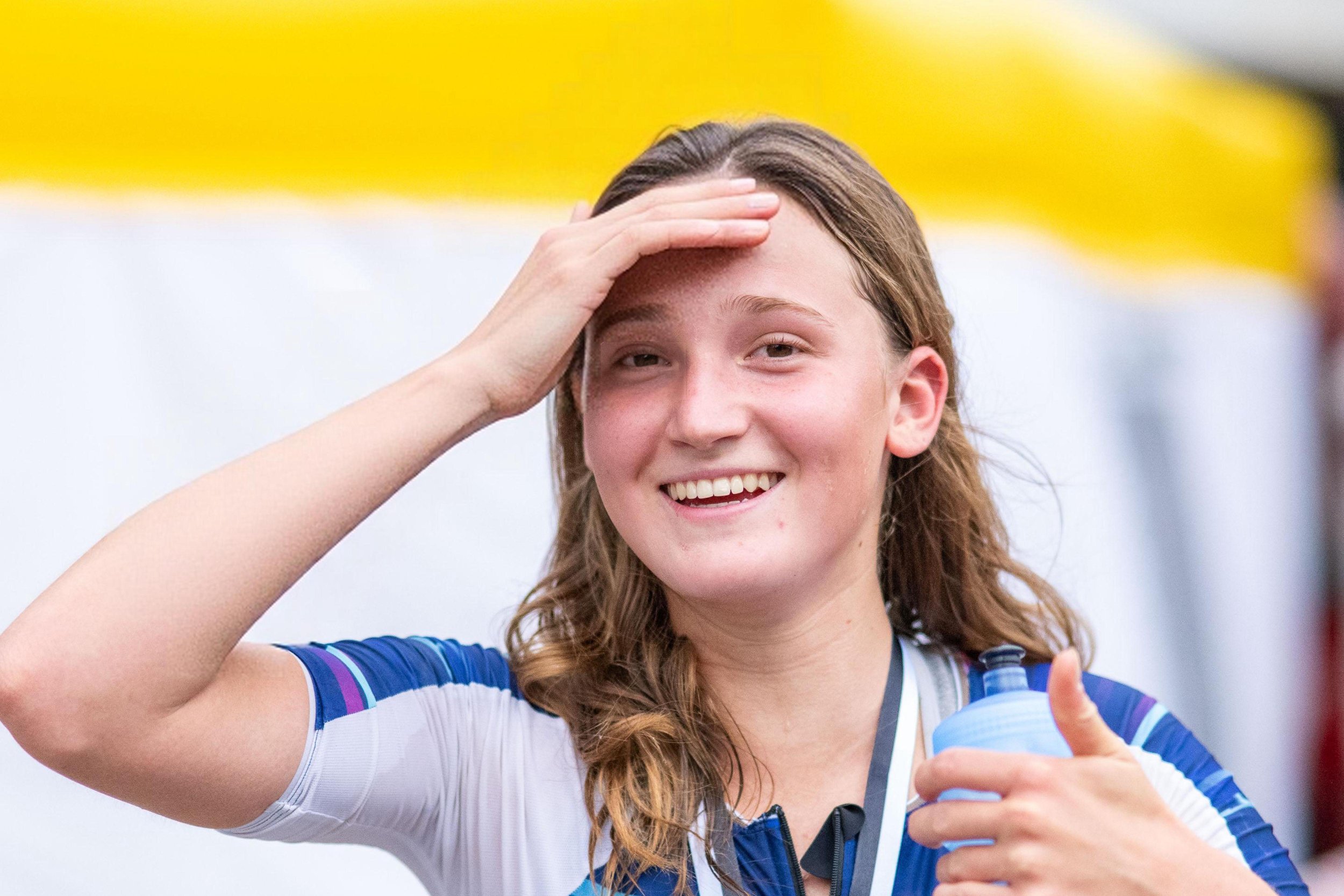 Junge Athletin nach einem Lauf, schwitzt, lächelt und hält eine Wasserflasche, während sie die Stirn mit der Hand berührt.