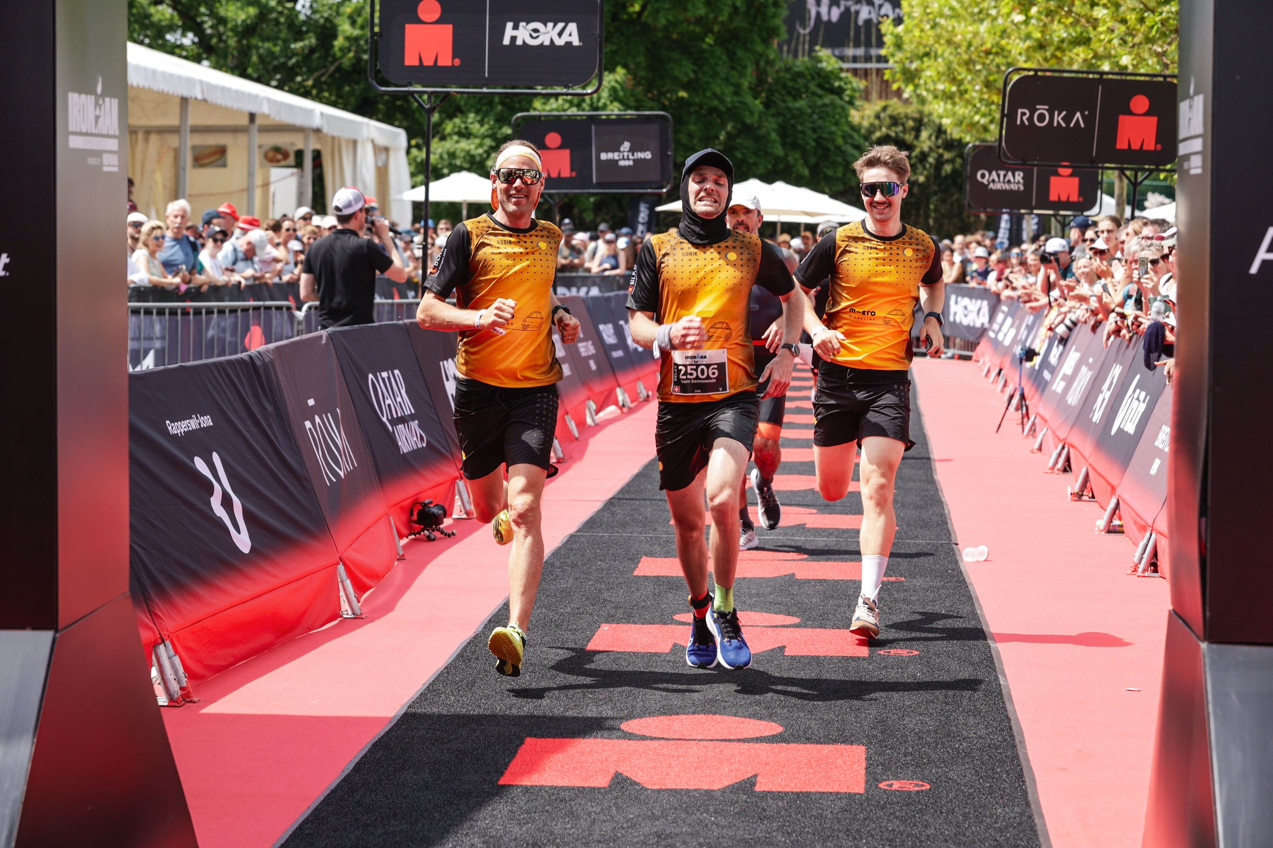Drei Läufer laufen gemeinsam die Ziellinie eines Marathonlaufs bei schönem Wetter, mit jubelnden Zuschauern an den Seiten und Werbetafeln im Hintergrund.