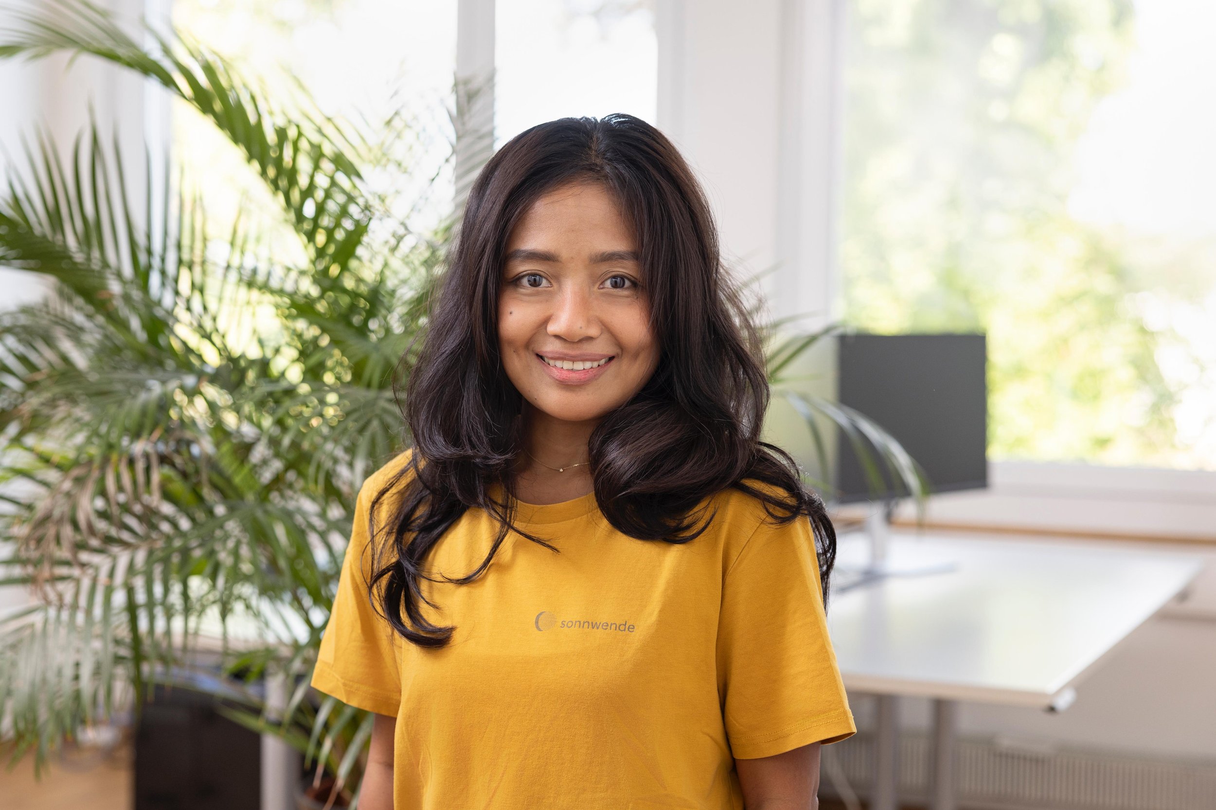Eine lächelnde Frau mit schwarzen, mittellangen Haaren, trägt ein gelbes T-Shirt mit der Aufschrift 'sonnwende', steht in einem hellen Raum mit großen Fenstern, umgeben von grünen Zimmerpflanzen.