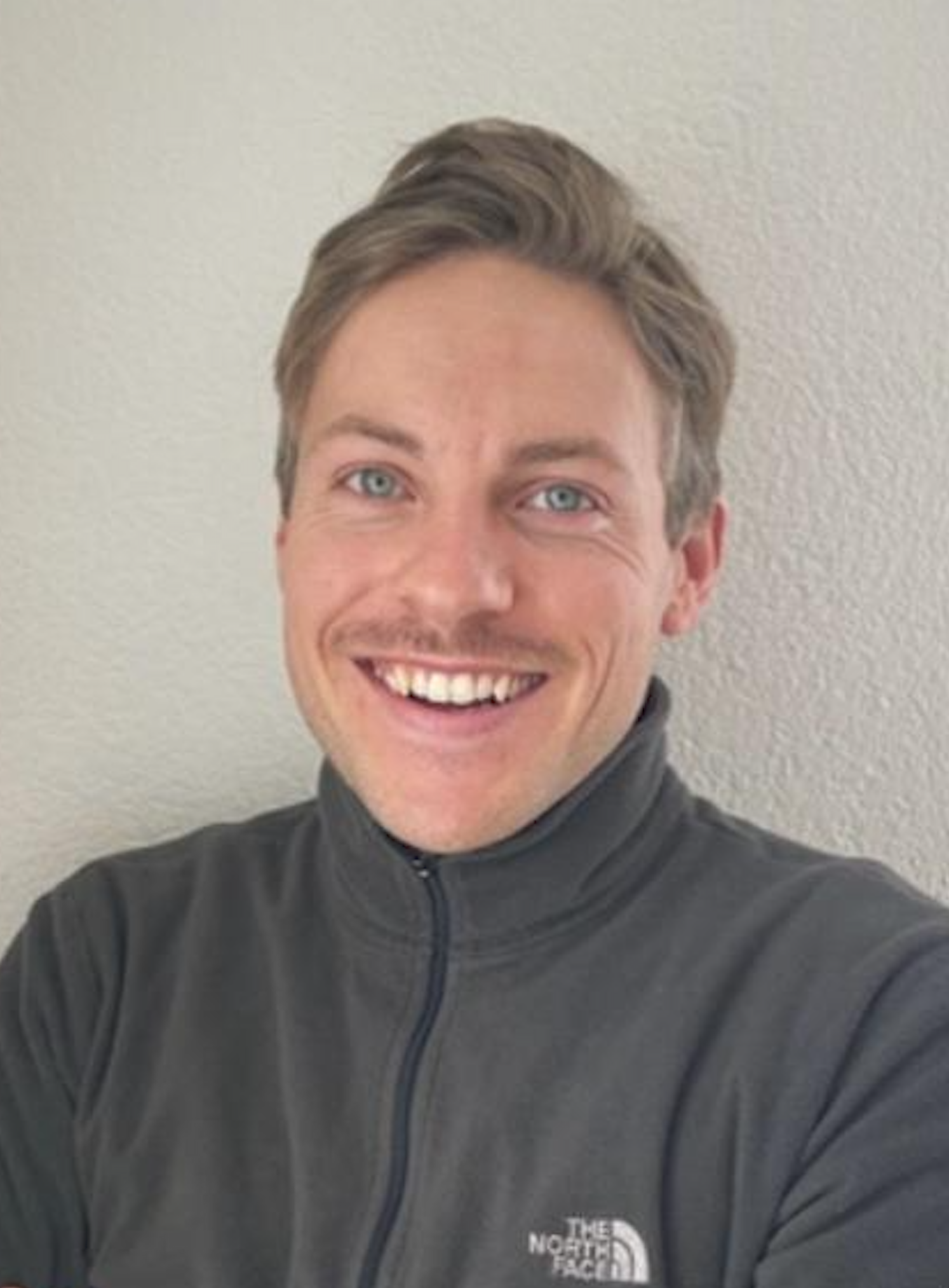 Ein Mann mit blauen Augen, braunen Haaren und einem breiten Lächeln trägt eine dunkelgraue Jacke mit Reißverschluss, auf einer hellen Wand.