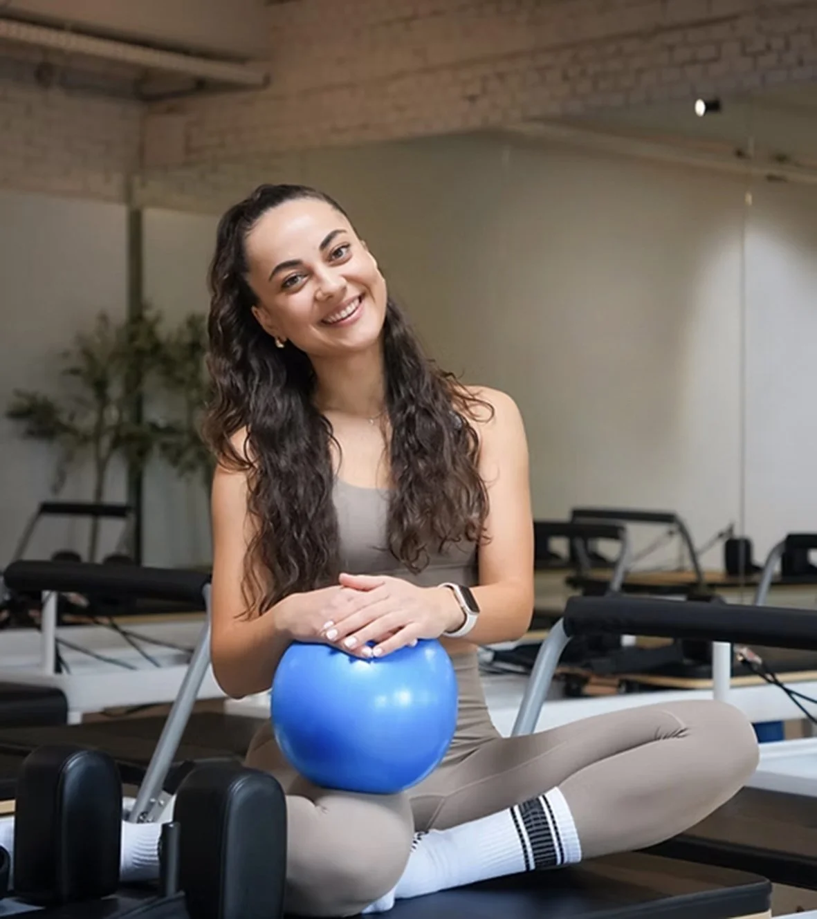 Een glimlachende jonge vrouw zit in een sportschool op een yogamat, met een blauwe oefenbal op haar schoot en fitnessapparatuur op de achtergrond.