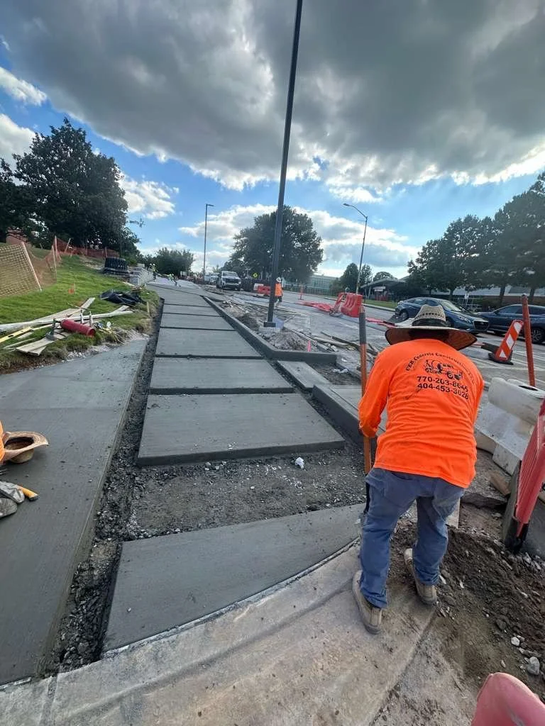 A construction worker in an orange shirt and hat working on a sidewalk under a cloudy sky.