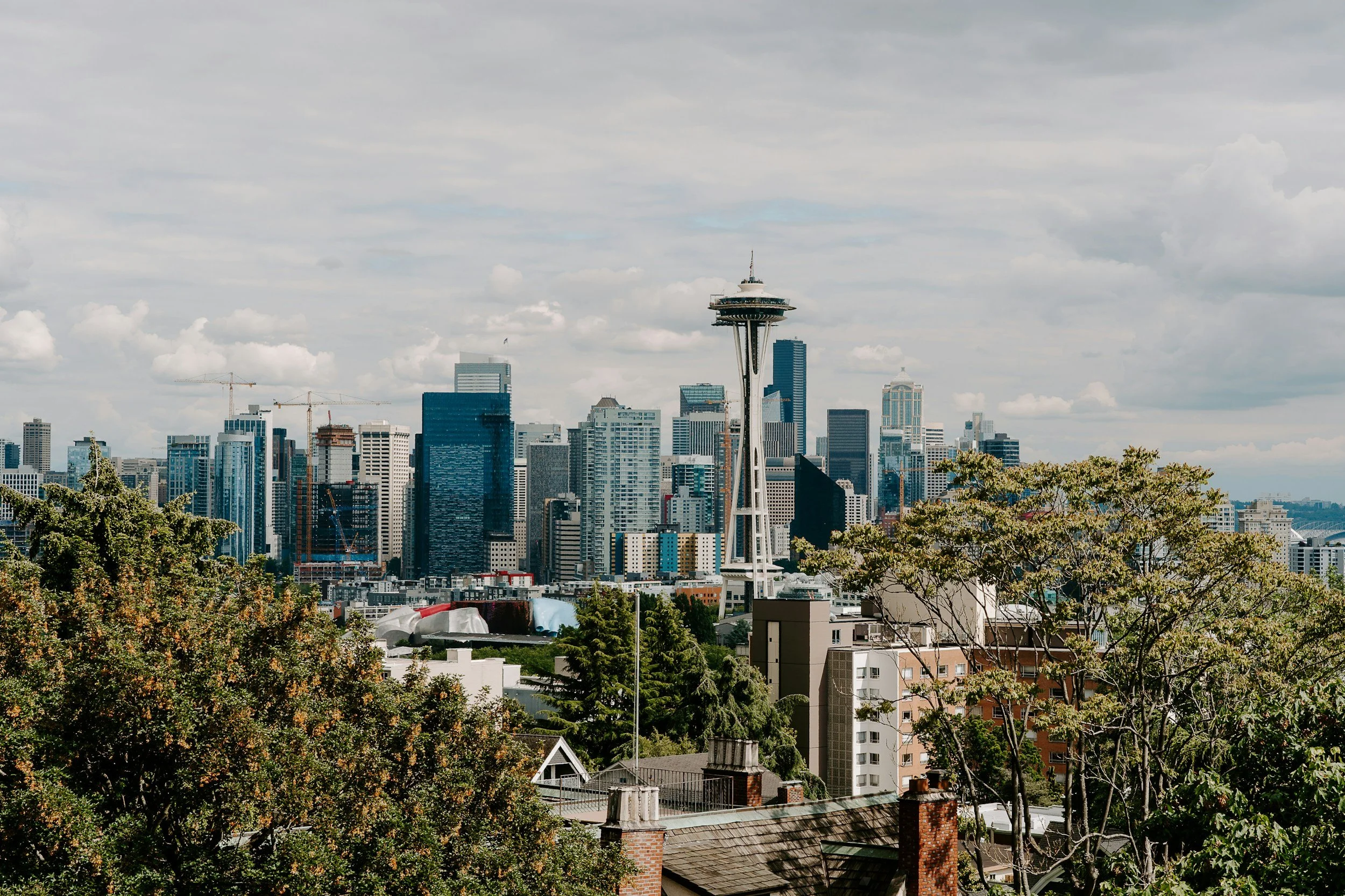 Seattle skyline featuring the Space Needle, with modern skyscrapers, some construction cranes, and a partly cloudy sky.