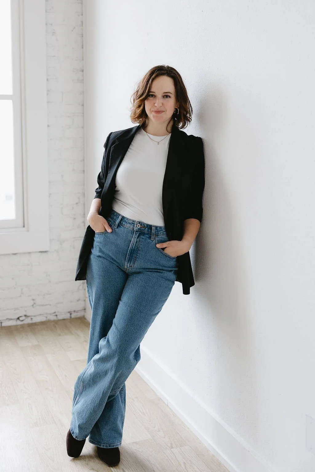 A young woman with shoulder-length wavy brown hair, wearing a black blazer, white t-shirt, blue jeans, and black shoes, standing in a bright room with a white brick wall and a window.