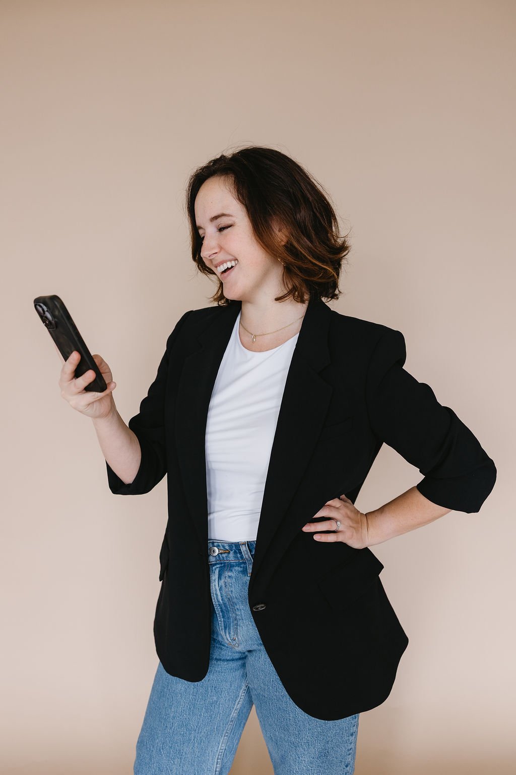 Chelsea Kern, smiling and looking at her phone against a plain light beige background, is a friendly young female real estate agent ready to assist clients in Seattle, Shorline, Bothell, Bellevue, and the surrounding Greater Seattle Area.