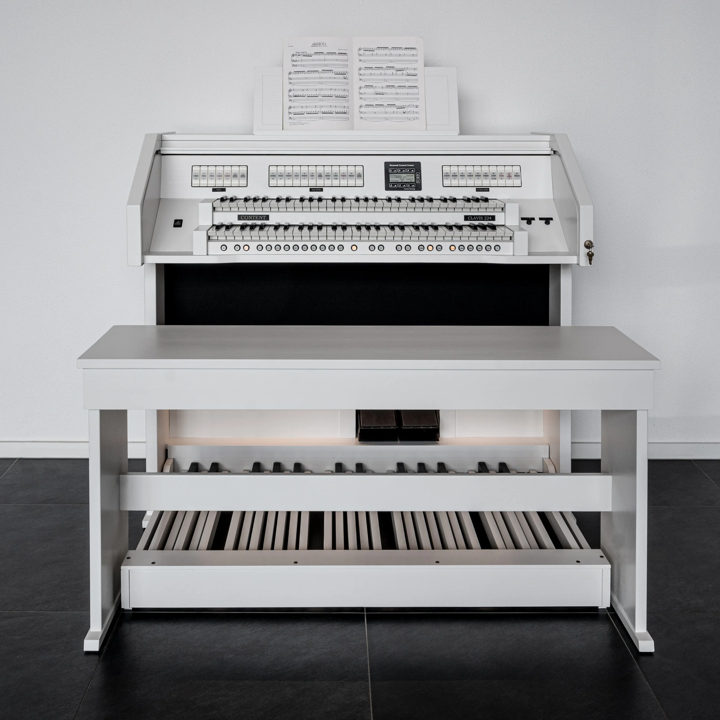 White electronic organ with sheet music on the stand, placed against a plain wall, on a black tiled floor, with a white bench in front.