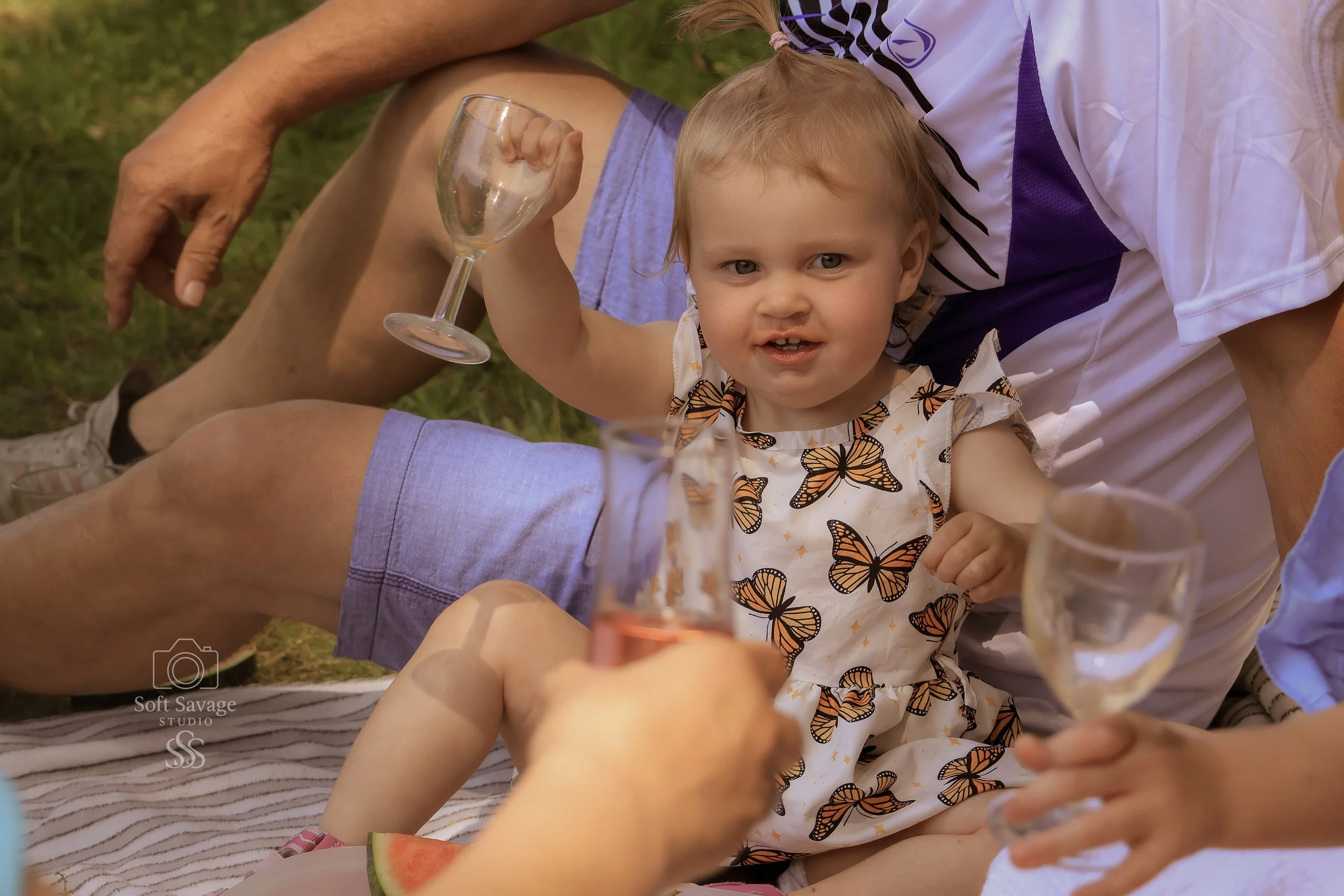 Ein kleines Mädchen mit Schmetterlingsmuster auf ihrem Kleid, das mit Erwachsenen bei einem Picknick im Freien sitzt, wobei alle Gläser mit Getränken in der Hand halten.