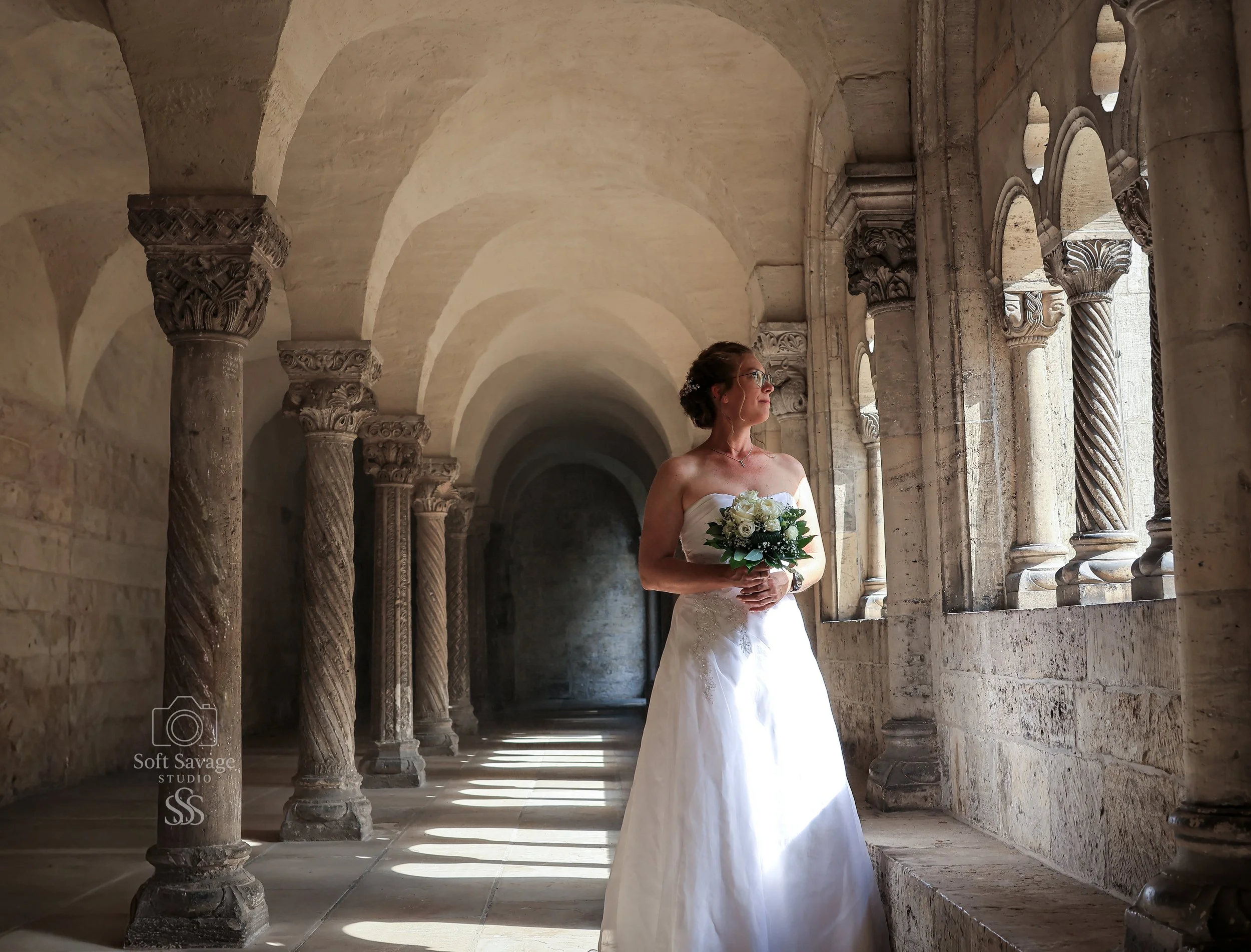 Braut in weißem Brautkleid mit Blumenstrauß steht in einer historischen, gotischen Kirche mit Steinarkaden und beleuchteten Fenstern, Blick nach rechts.