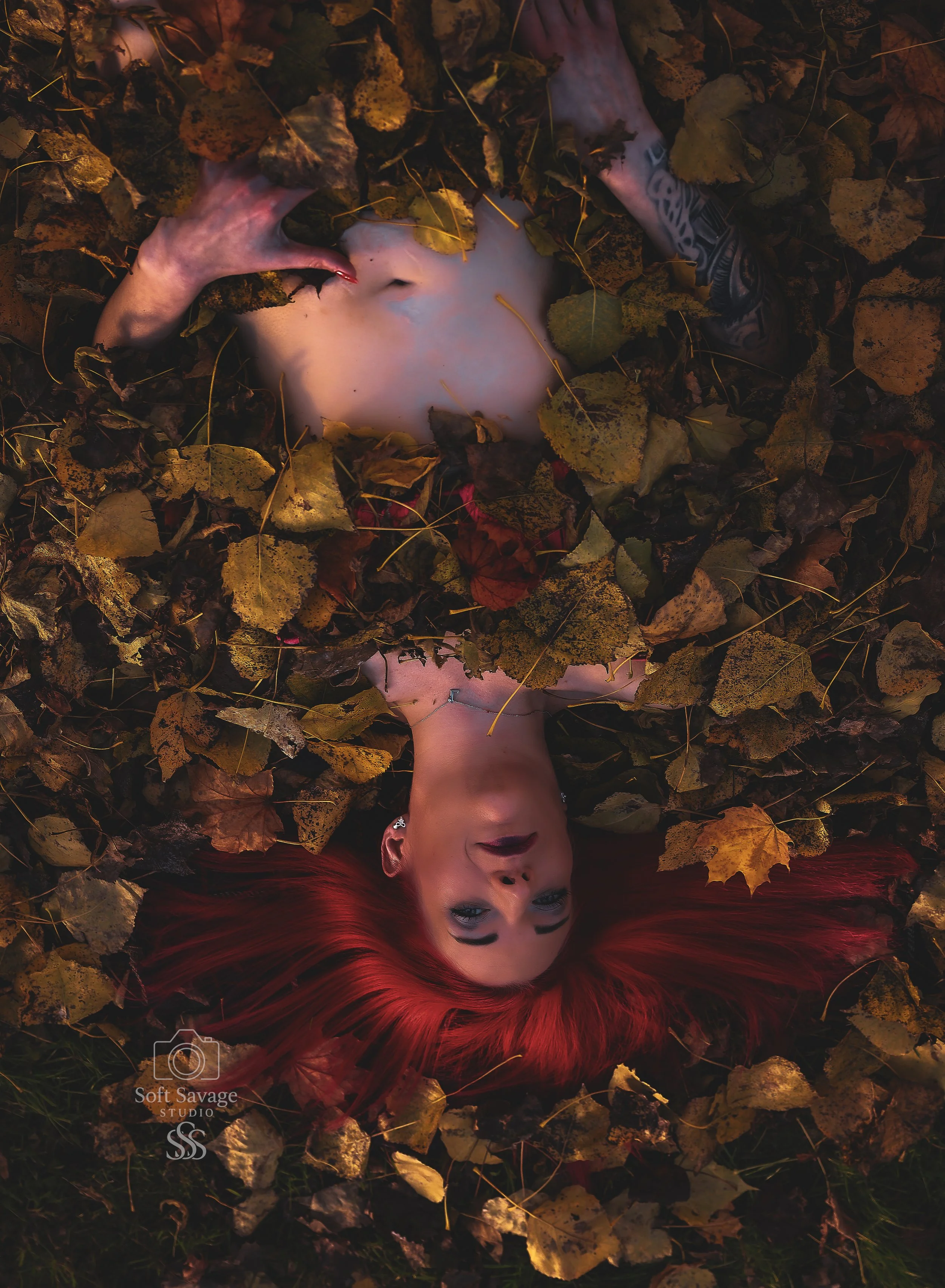 Eine Frau mit roten Haaren liegt nackt in einem Laubhaufen und blickt in die Kamera.