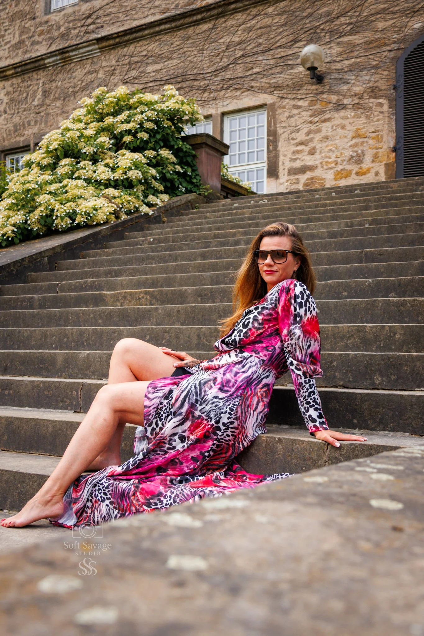 Frau in einem bunten, tierprintartigen Kleid sitzt auf Steinstufen vor einem alten Gebäude mit einer Blumenhecke.