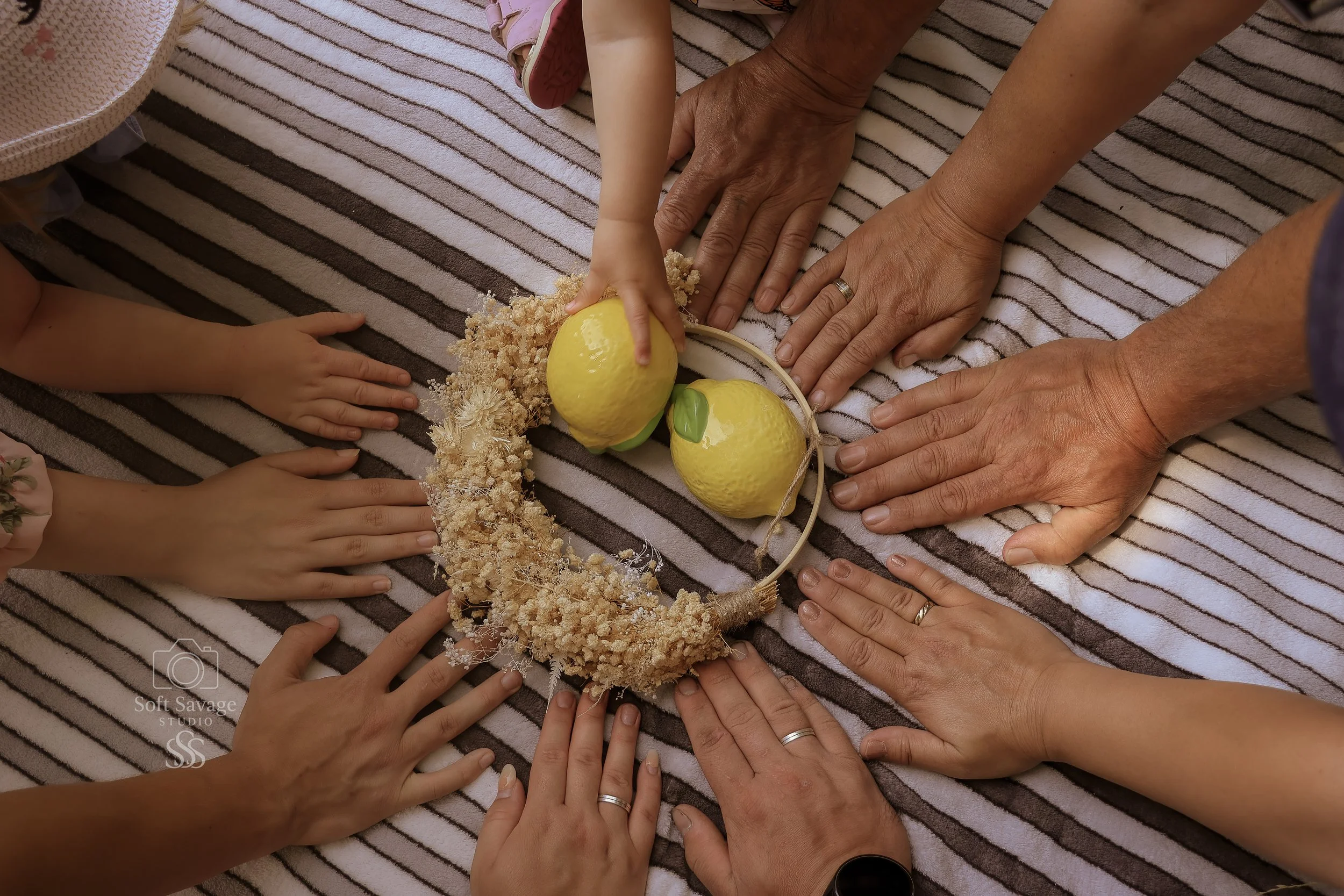 Mehrere Hände verschiedener Menschen, die um eine Schlafmaske mit Zitronenmotiv und Blumenkranz liegen, auf einem gestreiften Stoff. Zwei Kinderhände, drei Erwachsenehände, einige haben Ringe, die Hände sind in einer freundlichen Geste.