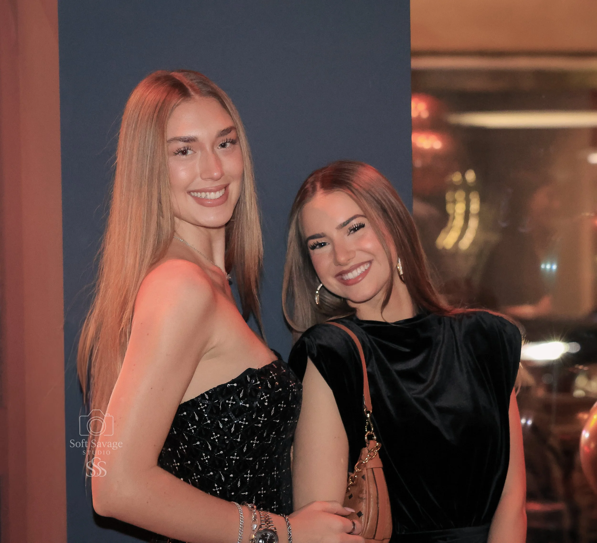 Zwei lachende Frauen bei einer Party in einem dunklen Raum, eine trägt ein schwarzes Kleid mit Spitze, die andere ein schwarzes Samtkleid mit großen Ohrringen.