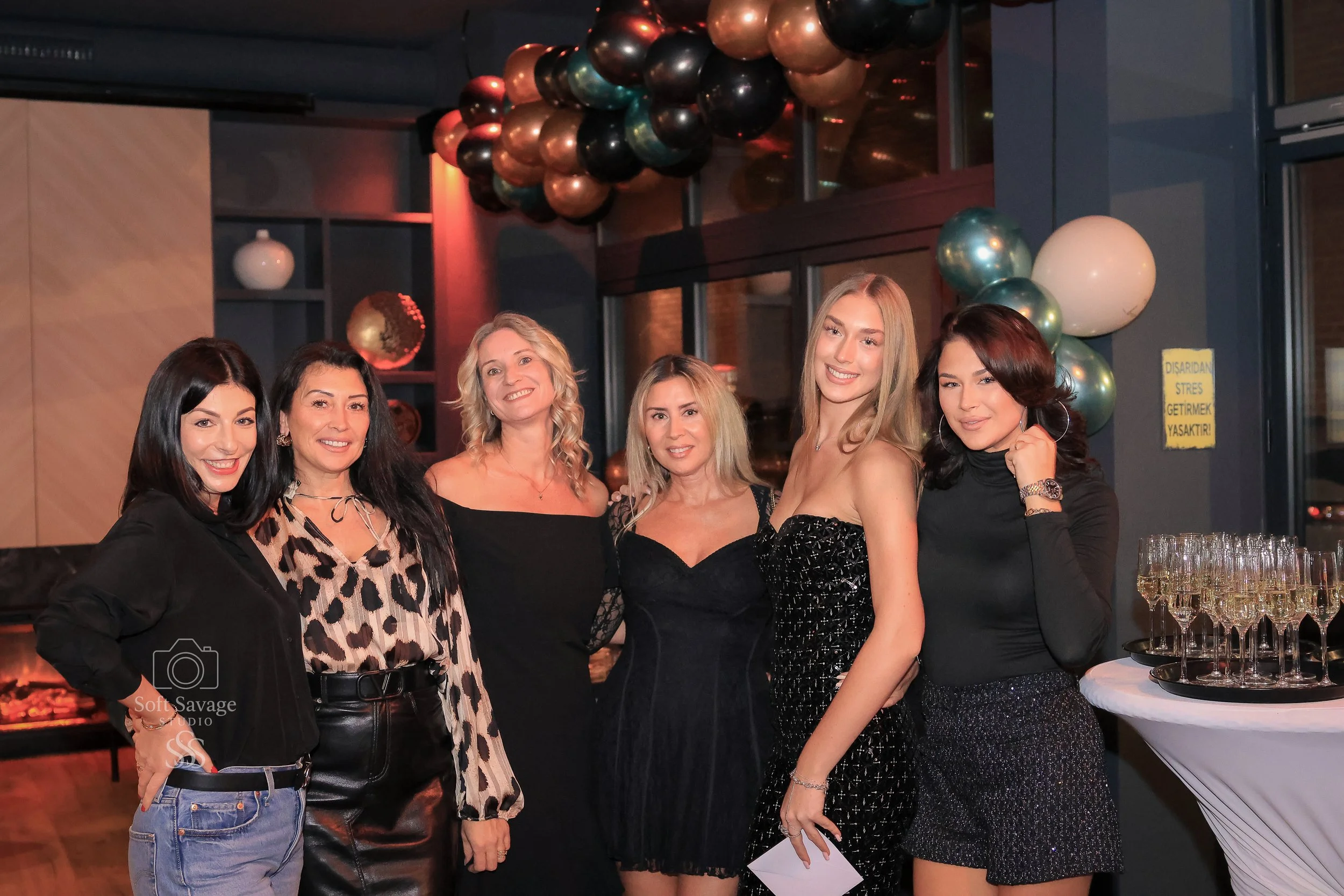 Sechs Frauen auf einer Party, mit bunten Ballons im Hintergrund, die lächeln und Fotos machen.