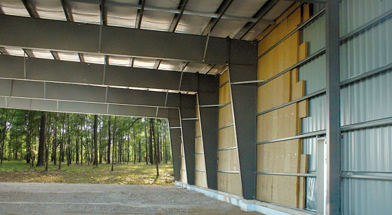 The interior of a building under construction, showing metal framing and a large open garage door with a view of a wooded area outside.