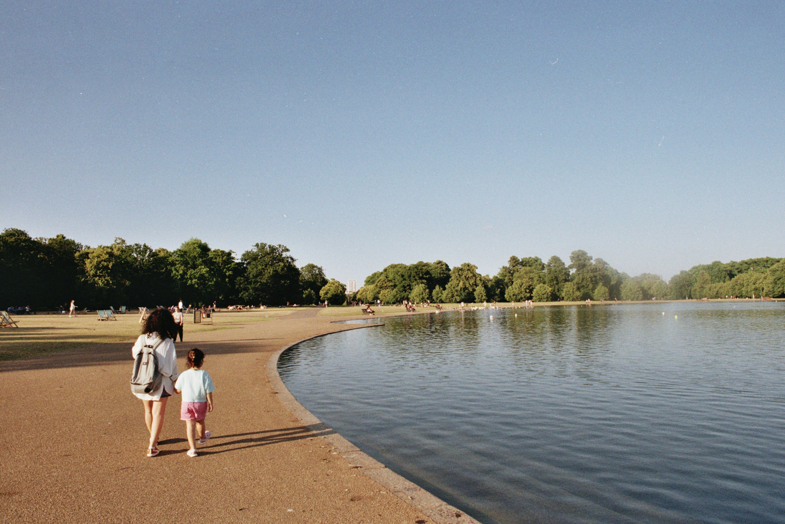 People walking along a curved lakeside pathway in a park with trees and a large body of water, under a clear blue sky.