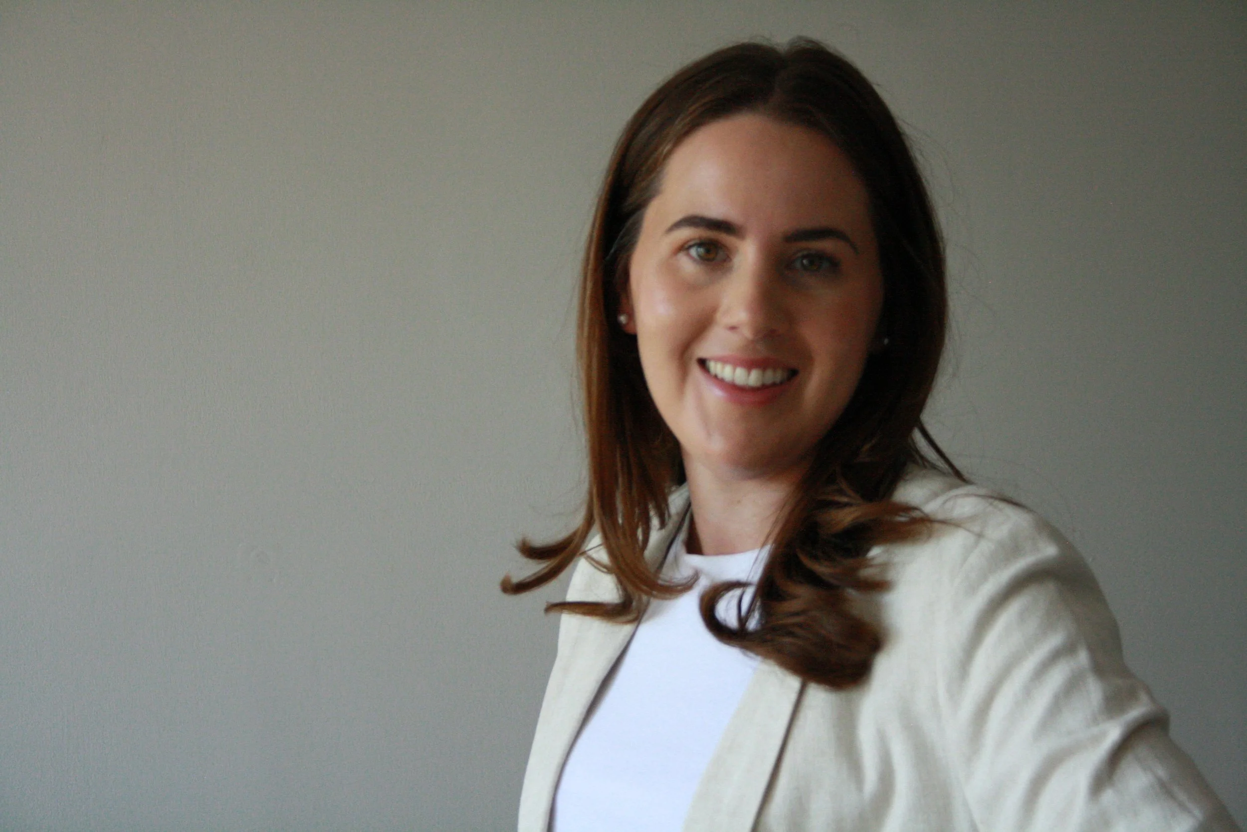 A woman with long brown hair, wearing a white shirt and beige blazer, smiling at the camera against a plain gray background.