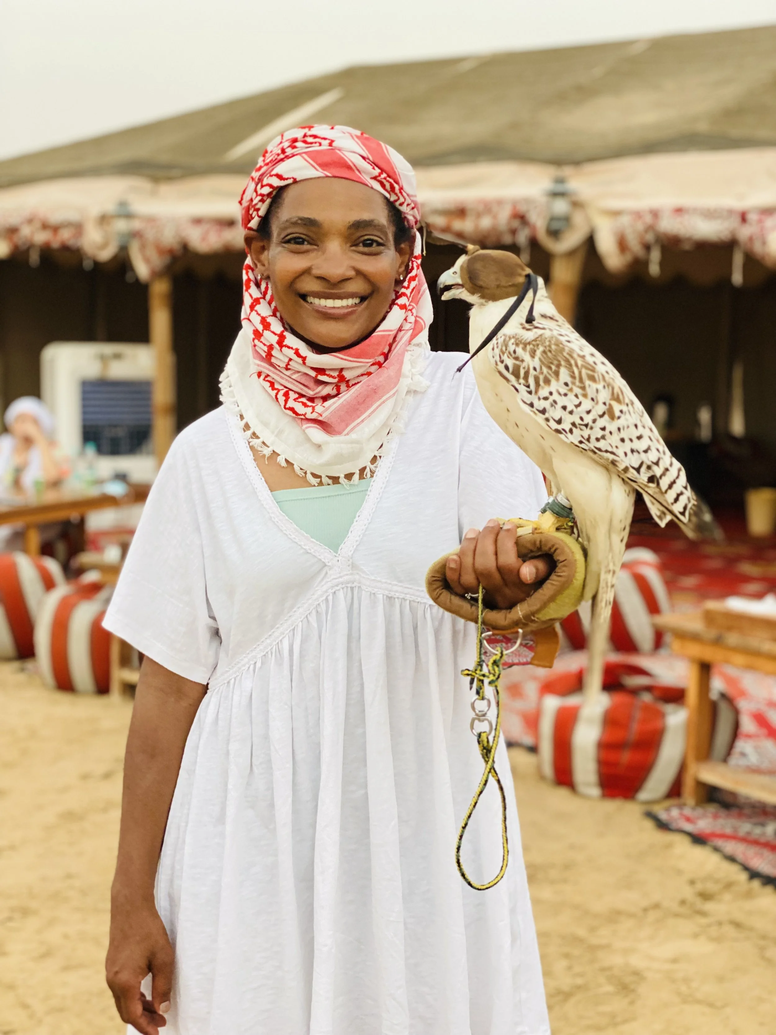 Woman with a red and white head scarf holding a falcon indoors, with textiles and seating in the background.
