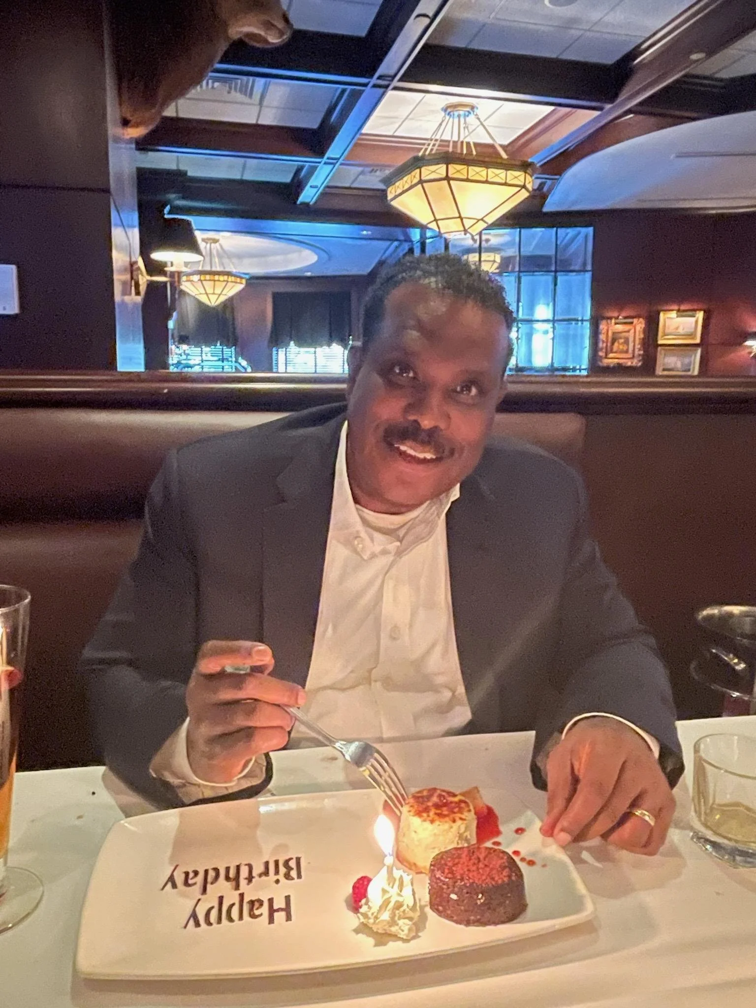 A man celebrating birthday at a restaurant with a lit sparkler on a dessert plate on a white rectangular plate with 'Happy Birthday' written on it, surrounded by chocolates, whipped cream, and berries.